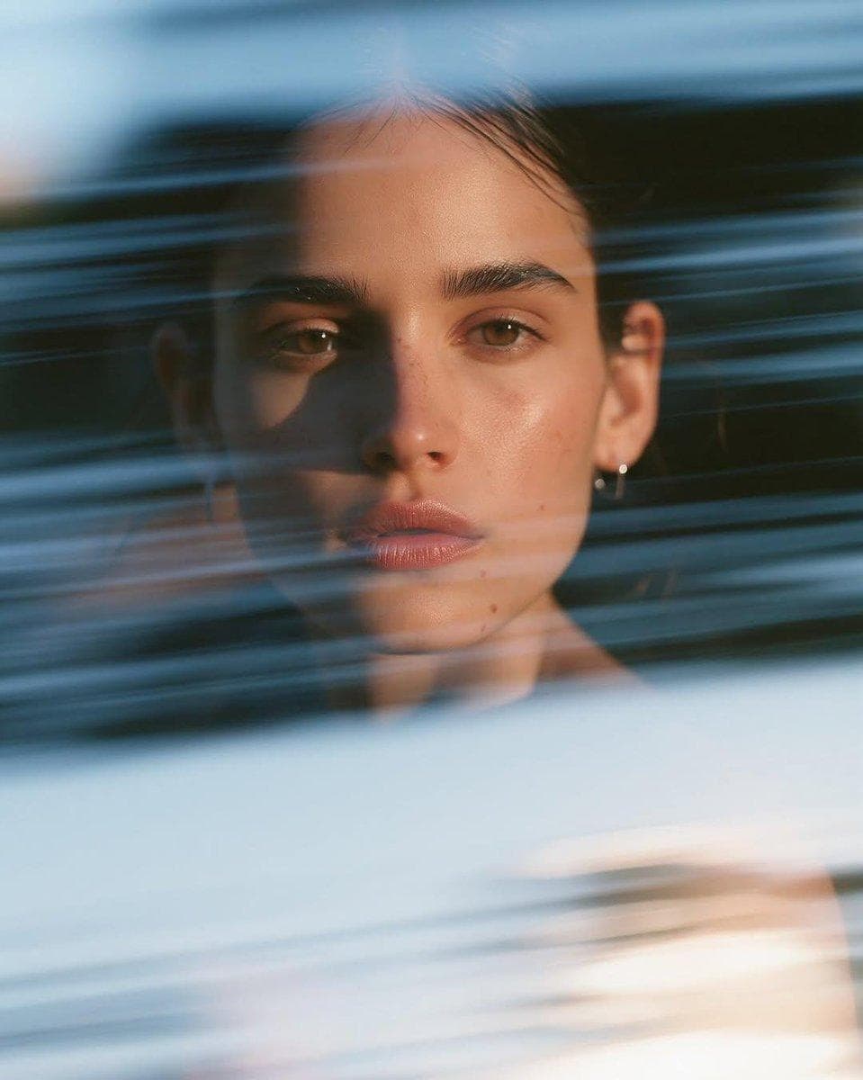 Ultra-realistic cinematic close-up portrait of a beautiful young woman with sleek dark hair pulled into a clean low bun, thick natural eyebrows, subtle freckles, glossy nude lips, flawless skin with realistic pores, wearing small silver hoop earrings and a minimalist black outfit. She gazes directly into the camera with a calm, introspective expression. Dynamic translucent blue acrylic panels or glass strips sweep horizontally across the foreground, creating layered motion blur and abstract light streaks while her face remains perfectly sharp. Soft reflections, refractions, and subtle lens distortion add depth. Moody dark studio background, cool blue highlights balanced with warm golden edge lighting, cinematic contrast, shallow depth of field, dreamy atmosphere…