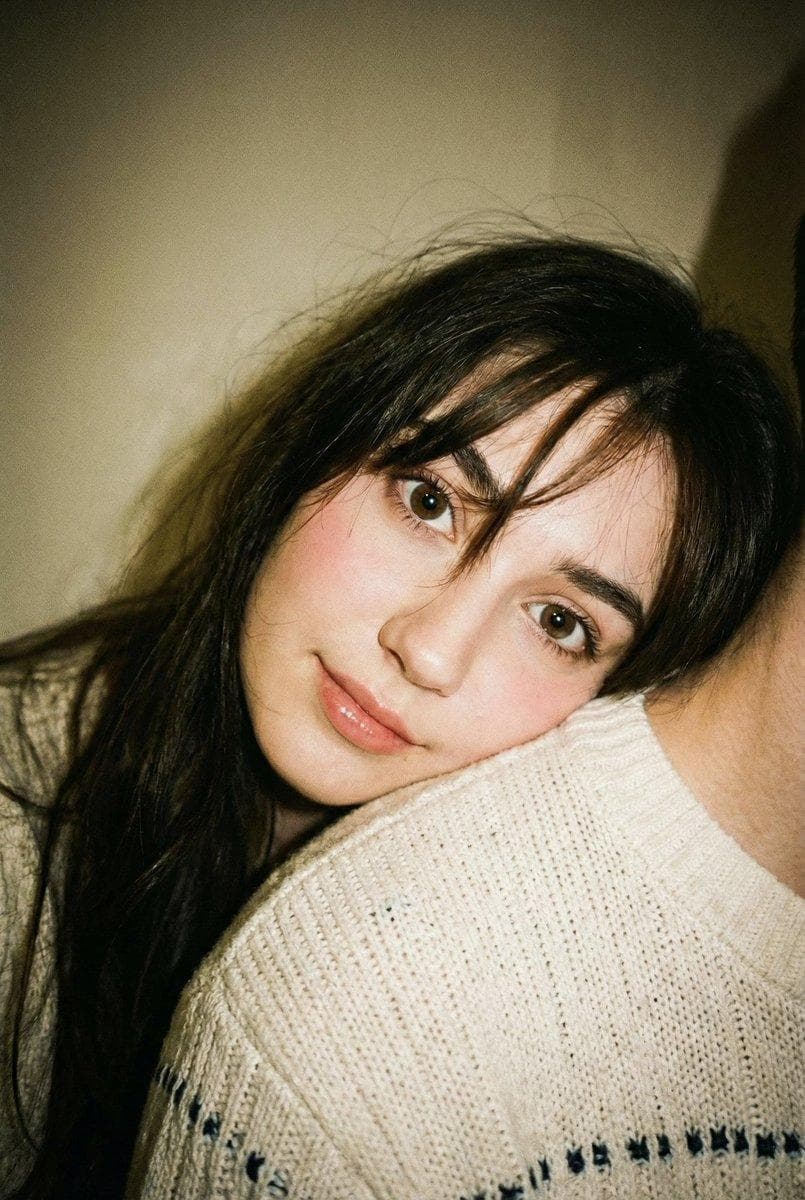 Edit this photo without changing the face. Portrait aspect ratio 2:3. Disposable camera aesthetic with a candid, warm, and slightly natural blur. The image should feel like a spontaneous moment captured on a single-use film camera, full of nostalgia, intimacy, and a cozy homey vibe. A young woman is leaning affectionately on her partner’s shoulder. Her face is turned toward the camera with her head slightly tilted. One hand presses her cheek, creating a cute pose. Innocent expression, big open eyes, with a soft smile or neutral lips. The partner is only partially visible (shoulder and neck only), creating a private, intimate moment. She is wearing a white or cream knit sweater with a simple design and small blue or black accents. Long black hair with natural bangs, slightly messy and not styled neatly. A few loose strands fall across the face for a raw, real film-photo look. Makeup is very minimal and fresh. Natural skin texture (not flawless or overly smooth). Thin, natural eyebrows. Soft pink blush. Natural pink lips with a slight gloss. Wearing brown contact lenses. Background is a home interior with plain walls. Warm indoor lighting, slightly uneven, with soft shadows and natural light noise typical of film. No professional lighting setup. Shot on a disposable camera…