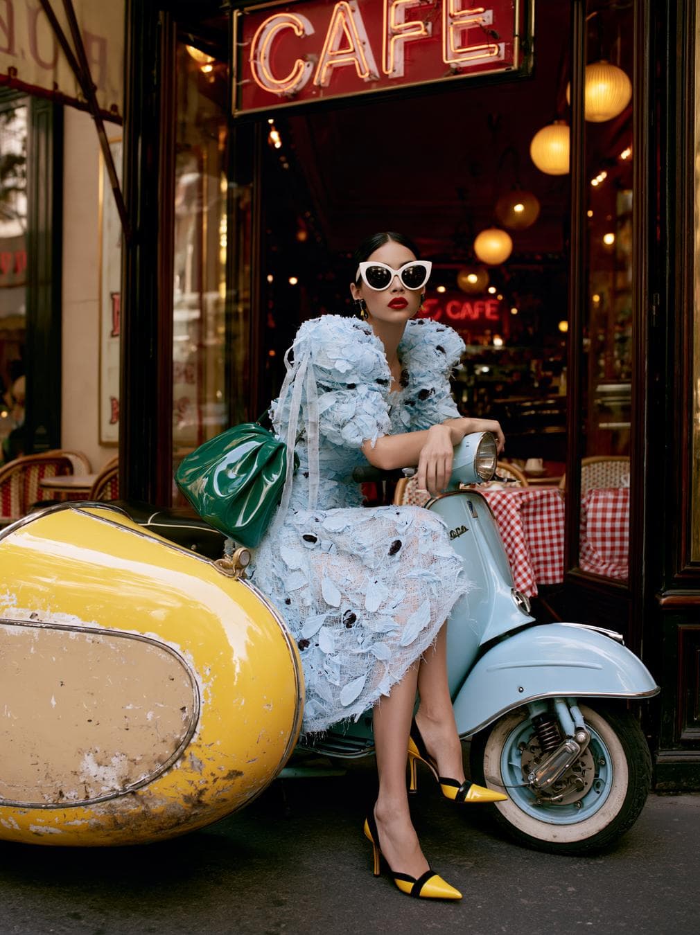 Ultra realistic high fashion street editorial, young adult woman outside a classic European café beside a vintage powder blue Vespa style scooter with an oversized glossy canary yellow sidecar. Eccentric, glamorous, slightly surreal 1960s Italian fashion fantasy meets modern editorial. Sleek jet black center parted low bun. Porcelain skin, sharp brows, dramatic black eyeliner, vivid matte red lips, oversized white cat eye sunglasses with black lenses. Pale icy blue couture midi dress: dense 3D ruffles, shredded tulle, feather like petals, organza strips, dark embellishments, voluminous cloud like silhouette, dramatic neckline, exaggerated shoulders. Glossy emerald green structured handbag, yellow pointed toe stiletto pumps with black accents. Seated elegantly on sidecar edge, composed aloof expression, full body editorial pose. Café: dark wood frames, vintage red "CAFE" sign…