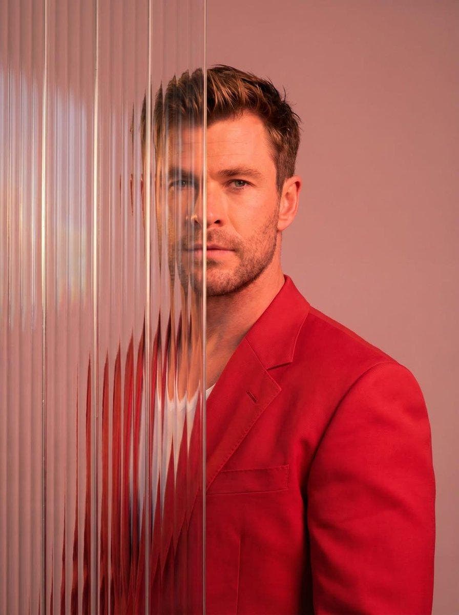 A striking editorial portrait photograph of a person wearing a red blazer, shot against a soft light pink studio background. The left half of the face and body is partially obscured by multiple overlapping vertical strips of tall narrow reeded ribbed frosted glass panels, creating a realistic distortion and refraction effect where the features appear blurred, shifted, and fragmented behind the glass. The right side of the face is clearly visible with sharp detail. Dramatic cool-toned studio lighting with a blue-purple color grade overall. The glass strips run vertically from top to…