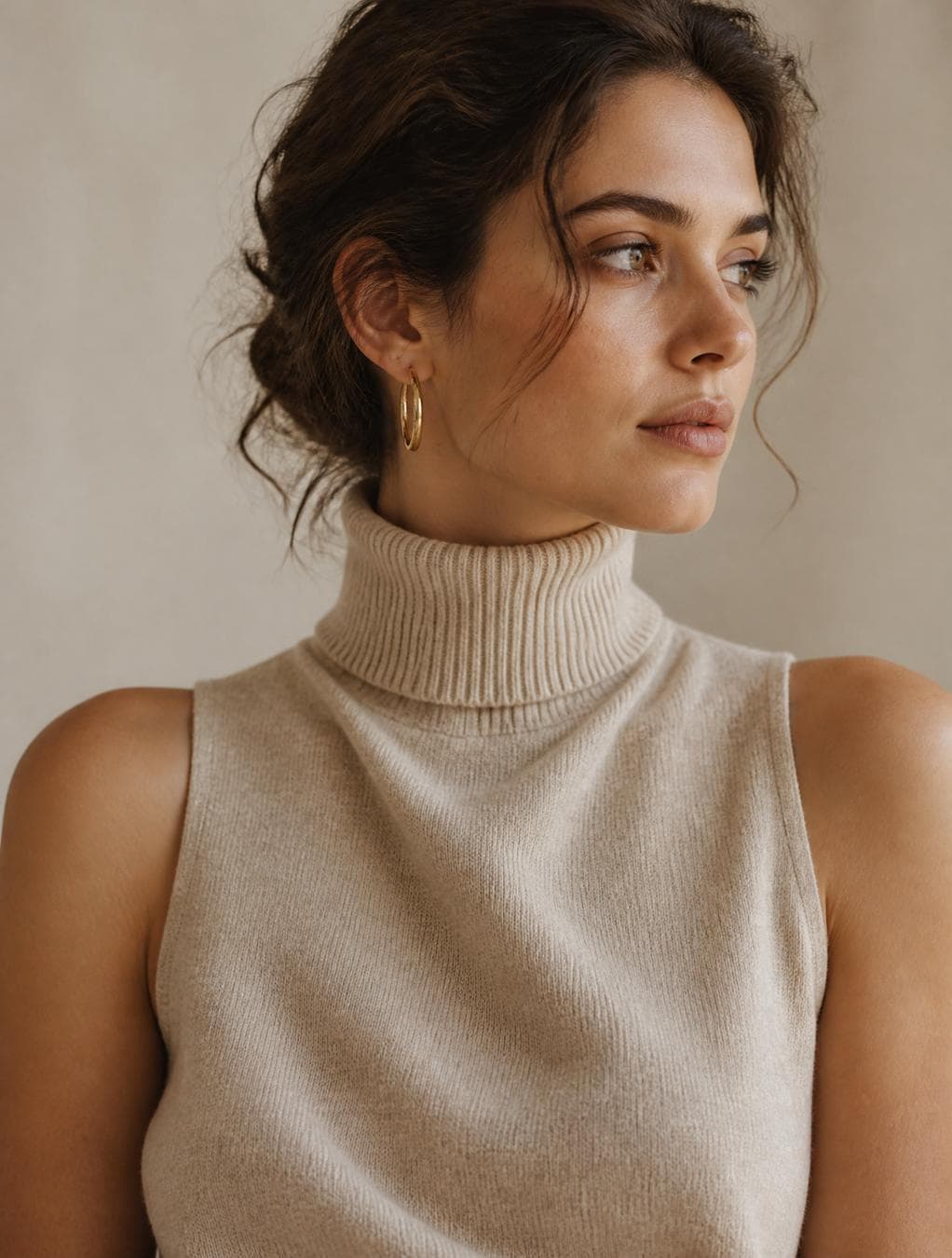 A single polished gold hoop earring catches a soft highlight on her left ear. She wears an elegant sleeveless knitted turtleneck sweater in a soft oatmeal beige color, crafted from premium cashmere and fine merino wool. The sweater features a high ribbed collar, refined knit texture, subtle woven fibers, and a perfectly tailored fit that gracefully follows her silhouette while leaving her…