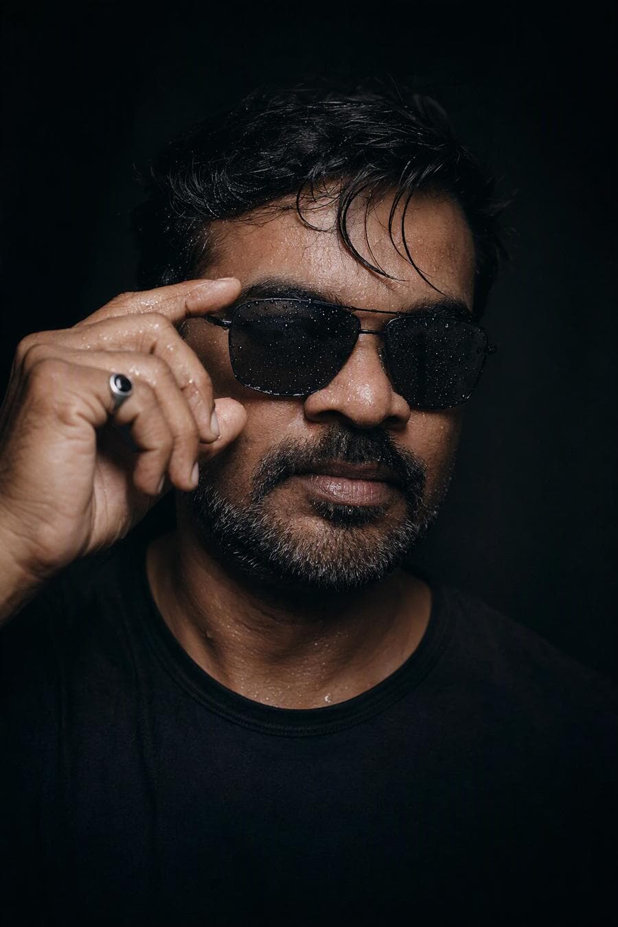 Ultra-realistic cinematic close-up portrait of a young man with wet skin and damp, messy hair. He is lightly touching the frame of dark, rectangular aviator sunglasses beaded with clear water droplets. The lighting is low-key and dramatic, creating deep shadows that emphasize the moisture on his skin and the fine texture of his beard. He wears a plain, form-fitting matte black t-shirt that blends into a dark, minimal background. Detailed hand gesture showing defined knuckles and a silver ring with stone on the…