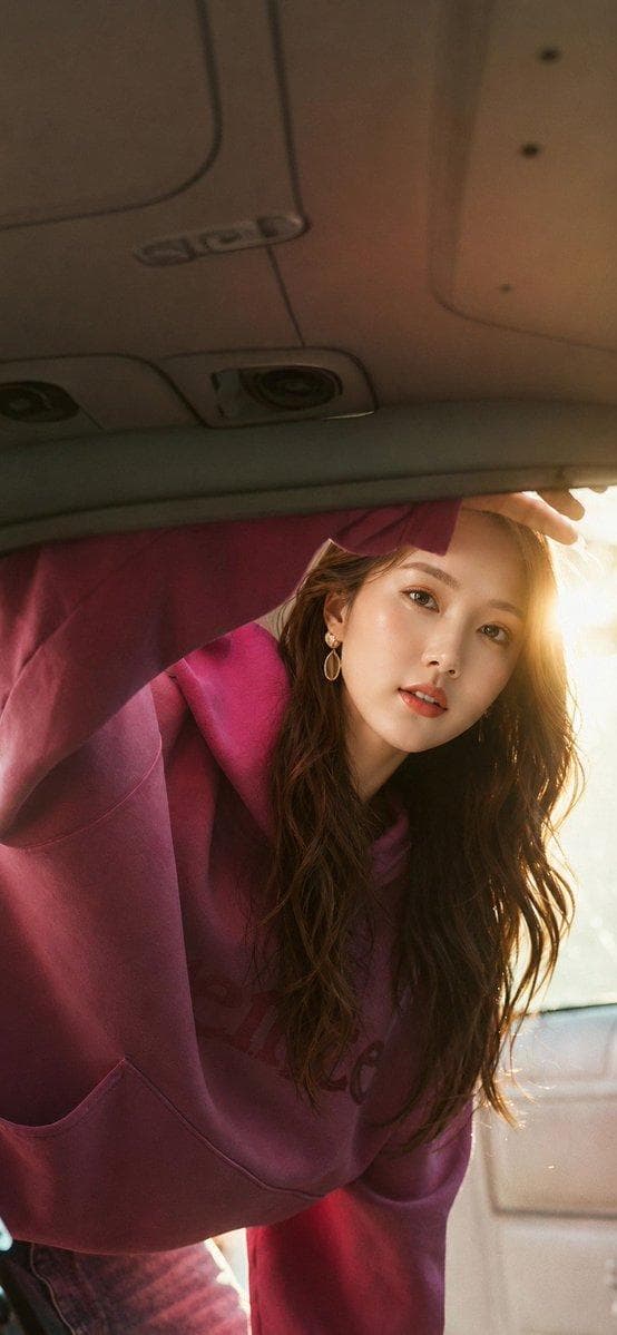 Ultra-realistic candid lifestyle portrait of a beautiful young beautiful woman [Face Reference]leaning into an open car trunk, viewed from inside the trunk looking outward. She wears an oversized deep magenta hoodie with a front pocket and subtle embossed logo, paired with matching casual bottoms. Her long, wavy dark brown hair falls naturally over her shoulders, with elegant gold teardrop earrings. One arm is raised above her head, resting on the trunk opening, while she gazes directly into the camera with a soft, calm, slightly curious expression. Golden hour sunlight streams from behind her on the right side, creating a warm rim light around her hair, soft lens flare, and cinematic glow. The car's interior roof, trunk frame, and overhead lights are visible in the foreground, naturally framing the composition. Warm earthy color grading, shallow depth of…