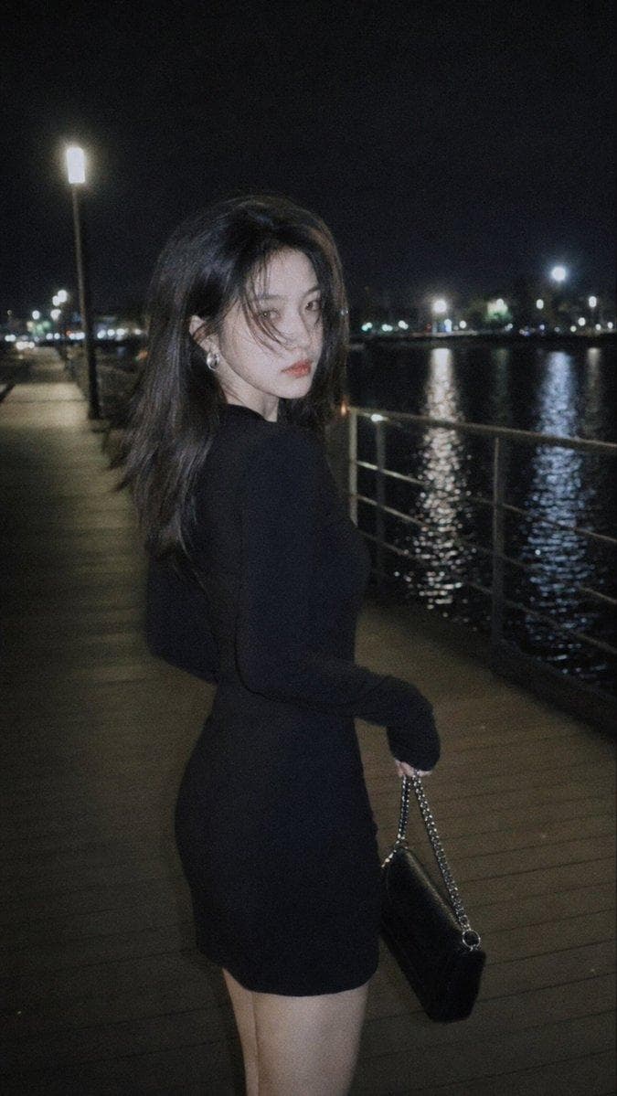 摄影风格：柔光CCD风 写真方向：夜色都市氛围街拍 场景方向：湖边夜间栈道 / 路灯 / 水面倒影 / 栏杆步道 服装方向：黑色长袖修身连衣裙 + 银色耳饰 + 小型链条包 气质标签：清冷、疏离、夜色感、克制、高级、真实抓拍感 五官方向：电影故事脸 身形方向：S型曲线 线条强调：中 镜头方向：大腿及上半身，近距离街拍构图 姿态动作：人物走在栈道边，听到摄影师喊声后回头看向镜头，一只手自然提包，身体带一点行走中的停顿感 光线氛围：夜晚路灯 + 水面反光 + 柔和闪光灯补光，人物前景被柔闪提亮，背景湖边夜色和路灯轻微虚化…