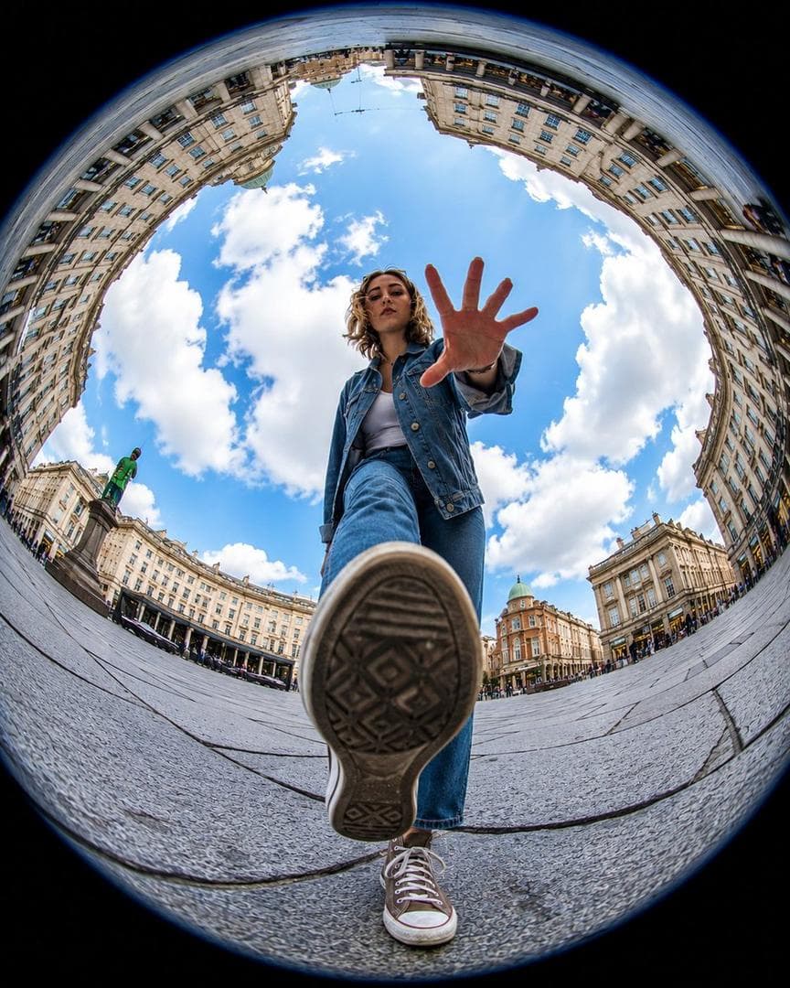 Ultra-realistic action photography, extreme fisheye lens, 8mm, worm’s-eye view from ground level, dynamic urban plaza scene. A stylish young person appears to be stepping over the camera, one sneaker sole dominating the foreground, hand reaching toward the lens.. Massive circular “tiny planet” perspective with historic European architecture curving around the edges of the frame, bright blue sky filled with fluffy white clouds at the center. Cinematic composition, dramatic distortion, ultra-wide angle, vibrant…