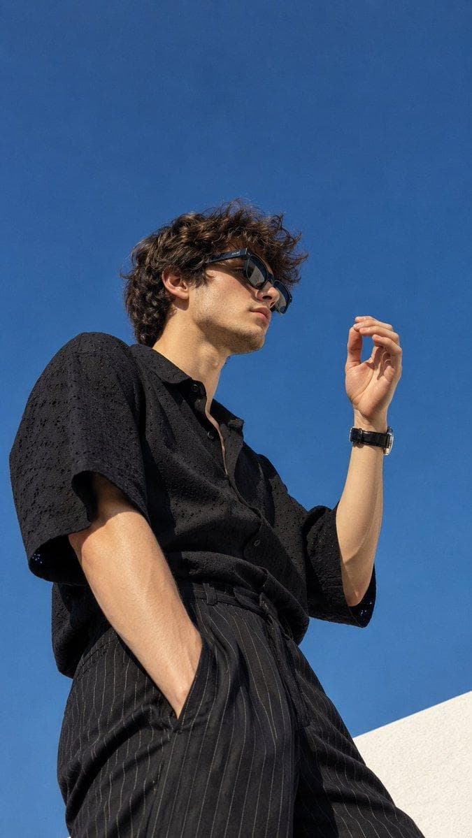 Create an ultra-realistic cinematic fashion editorial photograph of a young man captured from a dramatic low-angle perspective against a clear, deep cobalt-blue sky. He has medium-length, thick, naturally tousled dark brown hair with soft waves and delicate facial features. His face is shown in a side profile, looking slightly upward and toward the right side of the frame. He is wearing oversized futuristic black wraparound sunglasses with reflective lenses. He is dressed in a loose-fitting, textured black short-sleeve shirt with subtle eyelet embroidery and a relaxed, open collar. The shirt drapes naturally over his body. Pair it with high-waisted, relaxed-fit black trousers featuring thin, subtle vertical pinstripes. His pose is relaxed and stylish. His upper body is slightly angled away from the camera, one arm hanging naturally down in front of his body while the other arm is raised, with his hand positioned near his face in a gentle, elegant gesture. He wears a classic black wristwatch with a metallic case and a few minimal accessories. The photograph is taken from below, emphasizing the subject against the expansive blue sky. A small portion of a bright white architectural structure appears in the bottom-right corner. The composition is vertical, with the subject occupying the lower and middle sections of the frame and plenty of negative space…