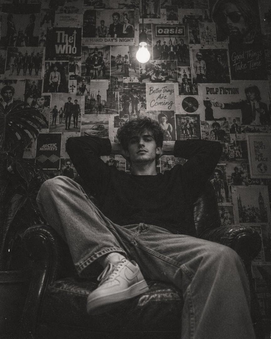 Create an ultra-realistic cinematic black-and-white editorial photograph of a young man relaxing deep inside a vintage dark leather armchair in a small, dimly lit bedroom or personal studio. Composition: Vertical portrait, 4:5 aspect ratio. Camera positioned slightly below eye level and directly facing the subject. The young man is centered in the frame, occupying the middle and lower portion of the image. His body is relaxed and naturally spread across the chair. Pose: The young man is sitting deeply and casually in the armchair, leaning backward into the seat. Both elbows are raised outward with both hands resting behind his head. His shoulders are relaxed. One leg is bent and raised toward the camera while the other leg extends naturally across the chair. His white sneakers are clearly visible in the foreground. The pose should feel effortless, relaxed, youthful and slightly rebellious. Subject: Young male with thick, naturally curly messy medium-length dark hair, slightly covering his forehead. Neutral, calm expression with a subtle serious look. Natural facial features, realistic skin texture, no exaggerated retouching. He is looking directly toward the camera. Clothing: Oversized plain black long-sleeve sweatshirt or loose black crewneck shirt, relaxed fit light-wash wide-leg denim jeans with natural wrinkles and folds, and clean white low-top sneakers. Clothing should look casual, slightly vintage and youthful. Environment: A dark vintage bedroom or creative personal room. The entire wall behind the subject is completely covered with an eclectic collage of old vintage magazine pages, retro advertisements, comic illustrations, fashion photographs, celebrity portraits, typography, posters and black-and-white printed artwork. The posters overlap each other naturally with uneven edges, creating a dense nostalgic wall collage. Lighting: A single exposed incandescent light bulb hangs from the ceiling near the upper center of the frame, directly behind and slightly above the subject. The bulb is glowing brightly and becomes the main practical light source. Strong falloff into deep shadows around the room. Soft light illuminates the subject's face and hair while leaving the edges of the room dark. Photography style: Authentic 1990s/early-2000s analog film photography aesthetic, gritty monochrome black-and-white, high contrast, deep blacks, soft highlights, visible fine film grain, subtle dust and film texture, slight halation around the…