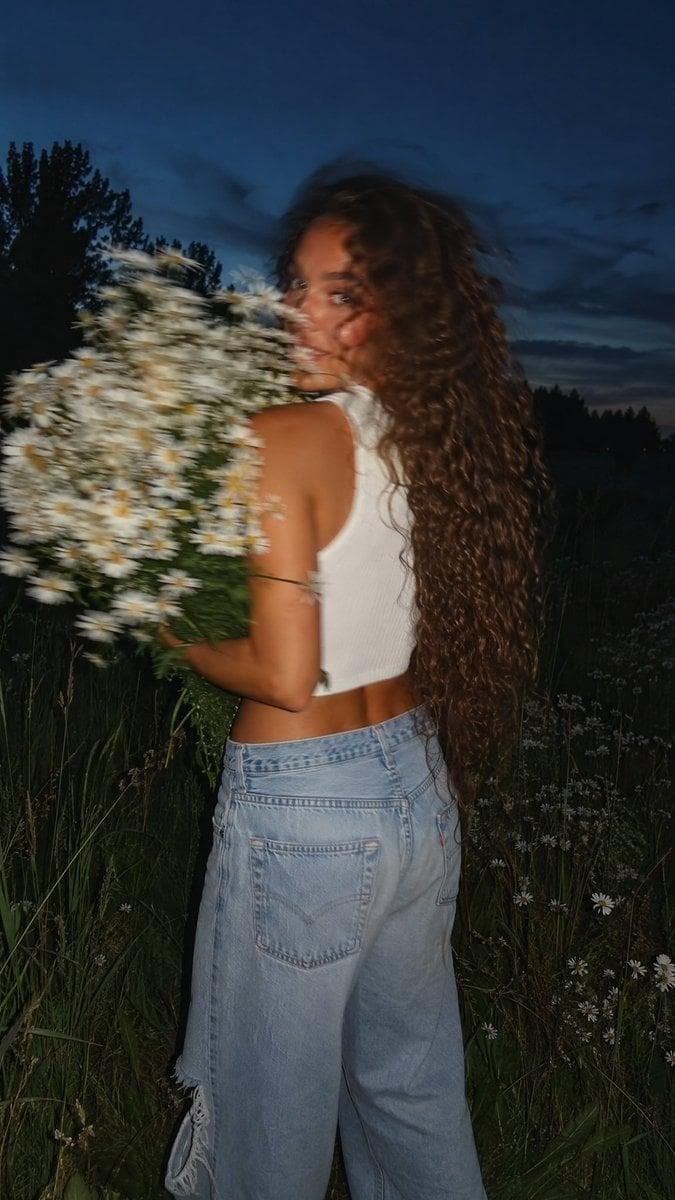 Ultra-realistic candid editorial photograph with a natural Canon G7X RAW aesthetic and subtle cinematic film grain. Vertical 9:16. A woman is captured in a fleeting, unposed moment while walking through a summer meadow at dusk. Seen mostly from behind, she suddenly glances back over her shoulder toward the camera, creating natural motion blur across her face, hair, and shoulders from the quick movement. Her expression feels warm, carefree, and genuine, as though the photograph was taken by chance. A massive bouquet of freshly picked white wild daisies is held close to her chest, partially covering the lower half of her face and upper body. She has exceptionally long, thick, naturally curly hair flowing well below her waist, with glossy, defined ringlets moving gently in the summer breeze. Individual curls remain visible despite the motion, adding depth and realism. She wears relaxed light-wash low-rise wide-leg jeans with subtle distressed details and a white lightweight knitted cropped sleeveless top featuring a high neckline, wide shoulder straps, and a fully covered back. The top has a soft premium knit texture, offering an elegant, modest summer look while remaining stylish and contemporary. The bouquet is oversized and luxurious, composed entirely of fresh white wild daisies with lush green stems, creating a premium countryside summer aesthetic. Her skin has a…