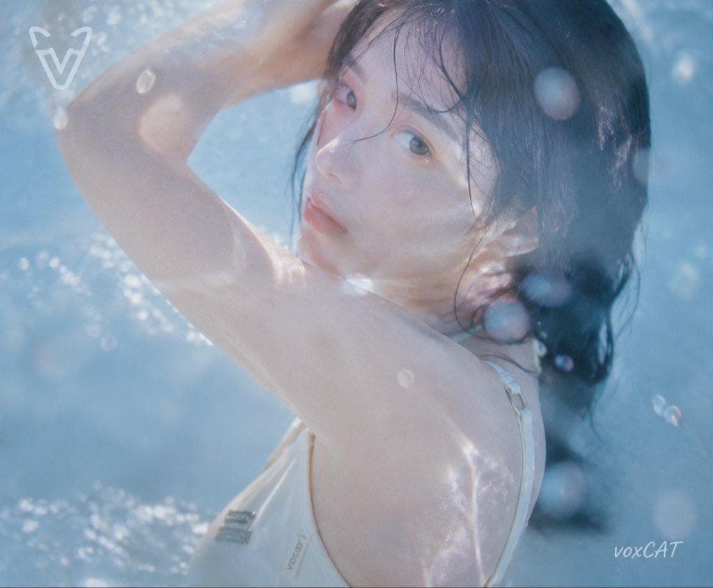 日系夏日水感写真，一位年轻女性角色，清透少女气质，湿润黑色长发，自然妆容，穿浅色简约泳装。 采用隔水拍摄视角，镜头位于水面或透明介质后方，前景出现水波折射、水雾、玻璃反光遮挡，人物被包裹在透明蓝色空间中。 人物半身动态构图，手臂抬起形成自然弧线，身体轻微转动，湿发贴脸，像夏日电影中的瞬间抓拍。 泳池蓝色环境，下午自然光，冷蓝水光包围人物，暖肤色，高曝光，低饱和，柔焦，朦胧梦幻质感，日系青春电影截图，老DV录像感，水下摄影效果。 整体加入VOXCAT品牌元素：…