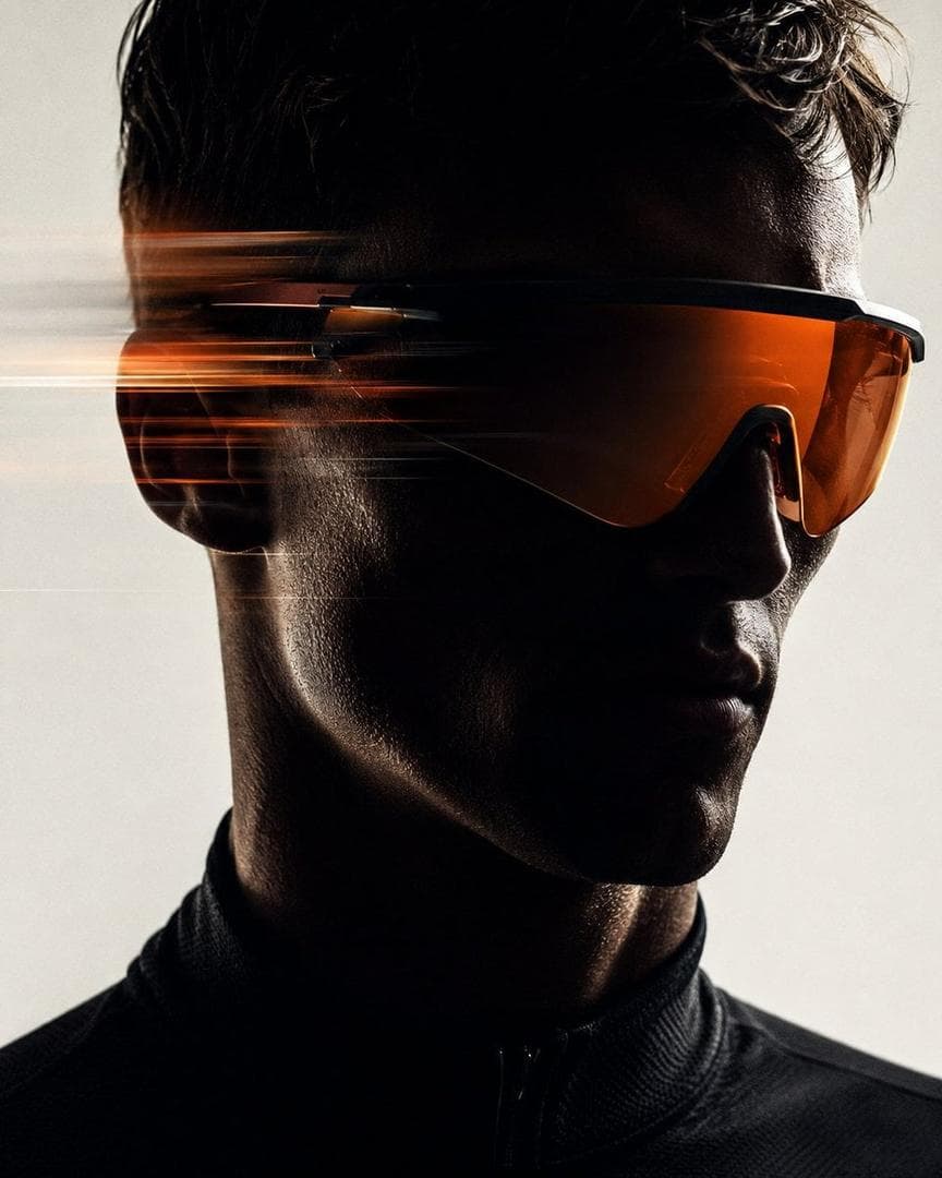 Futuristic high fashion sports editorial portrait of a male model wearing sleek oversized orange tinted visor goggles, close up head and shoulders composition, face mostly hidden in deep shadow, sharp jawline silhouette, dramatic low key studio lighting, clean off white background, horizontal speed motion blur streaks passing across the left side of the face and visor, premium athletic technology campaign aesthetic, minimal composition, cinematic contrast, grainy film texture, intense focused mood, ultra realistic photography, high end magazine…