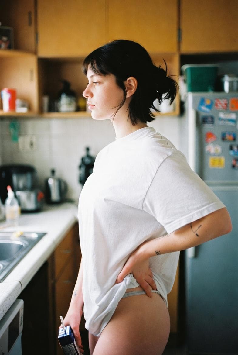 { "type": "image_generation_prompt", "style": "photorealistic, lifestyle photography", "sexualization": "none", "description": { "subject": "A young woman standing casually in a home kitchen, seen in side profile", "pose": "Relaxed posture, one hand resting on her hip while the other holds a small drink carton with a straw", "expression": "Neutral, thoughtful, slightly distant gaze looking forward", "appearance": { "hair": "Dark hair tied in a loose low bun with natural strands framing the face", "clothing": "Oversized white t-shirt worn casually, simple underwear partially visible due to the relaxed fit of the shirt", "tattoos": "Small, minimal black-ink tattoos on the forearm, subtle and understated" } }, "environment": { "location": "A modest, lived-in home kitchen", "details": "Wooden cabinets, countertop appliances, a refrigerator covered with colorful magnets and souvenirs", "lighting": "Soft natural daylight coming from a window, warm and slightly muted", "atmosphere": "Quiet, everyday domestic moment" }, "camera": { "angle": "Side profile view", "framing": "Medium shot capturing the subject from mid-thigh to head", "lens": "35mm lifestyle lens"…