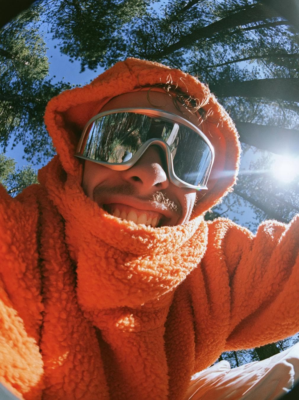 ULTRA-REALISTIC EDITORIAL PORTRAIT FROM INPUT PHOTO. VIBE: Y2K skate magazine cover, gorpcore outdoor streetwear, hyper-energetic. ENVIRONMENT: Dense pine forest looking up at a bright blue sky. LIGHTING: Harsh bright sunlight piercing through the trees, heavy lens flare, punchy daylight. STYLING (MALE): Thick orange sherpa fleece pullover, the hood/balaclava pulled tight around the face, aerodynamic silver reflective sports sunglasses. EXPRESSION: A wide smile close to the lens, Huge manic smile showing teeth, raw unhinged energy. the face takes up most of the frame. TECH…