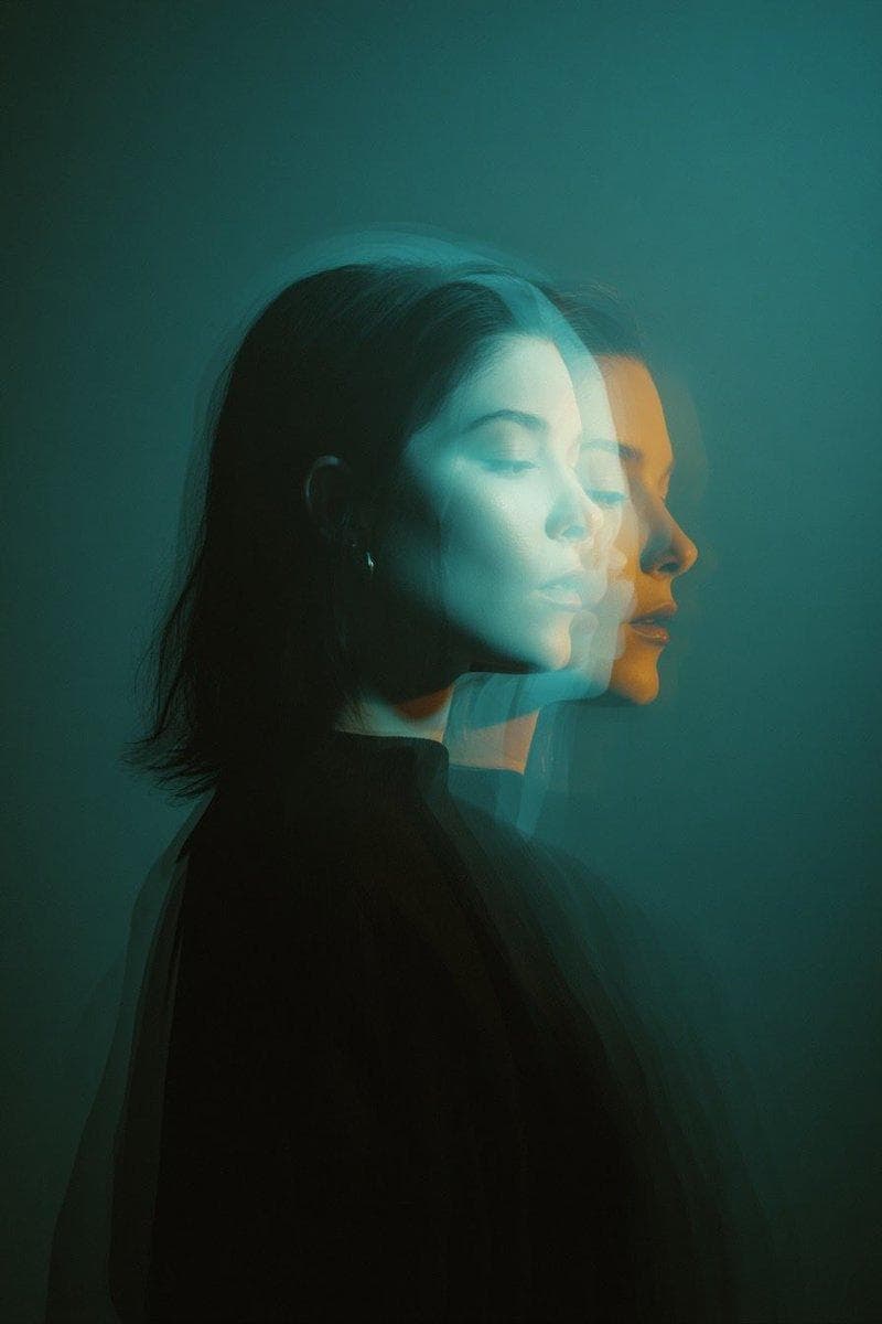 Cinematic double exposure portrait of a woman in side profile, two overlapping faces slightly offset, teal and orange moody lighting, soft motion blur, dark minimal background, high contrast, studio photography, dreamy atmosphere…