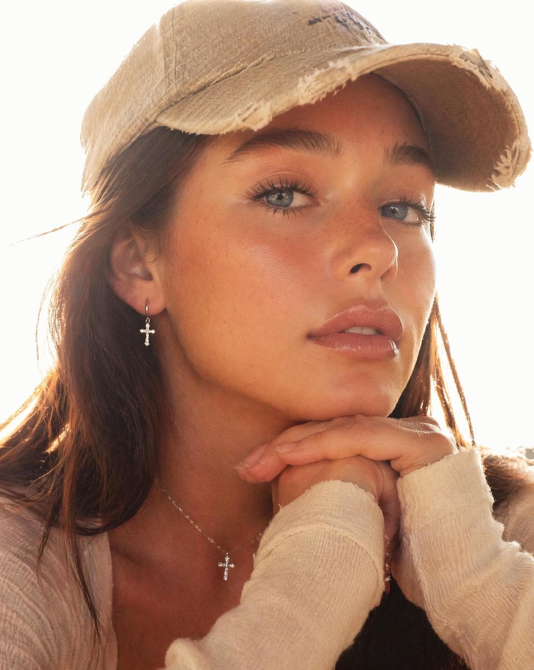 A young woman with long, dark hair wears a distressed beige baseball cap, her face tilted upward in a contemplative expression as she rests her chin on her interlocked hands. The composition is an intimate close-up, emphasizing the soft, natural skin texture and subtle subsurface scattering across her cheeks. Intense, overexposed backlighting creates a bright halo effect around her silhouette, washing out the background into a high-key, ethereal glow. The lighting is diffused and flat, minimizing harsh shadows while accentuating the delicate features of her face and the soft cotton texture of her long-sleeved top. Her gaze is directed slightly off-camera, adding a candid, spontaneous quality to the portrait, while the overall color palette remains warm, bright…