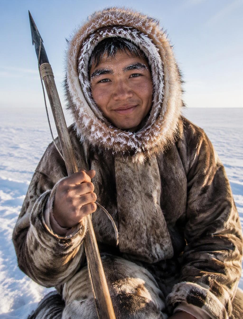 An ultra-realistic documentary portrait of a young Inuit hunter kneeling on sea ice, a gentle proud smile, frost on his eyebrows and the fur trim of his hood, holding a traditional carved harpoon, endless white tundra stretching behind. Tight vertical crop, bright cold Arctic daylight, rich…