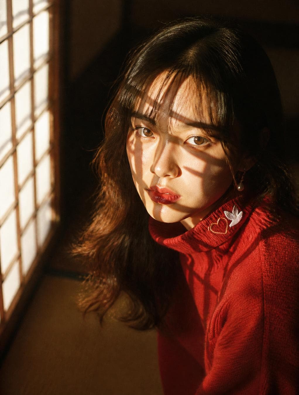 Intimate close-up portrait of a young Japanese woman kneeling near a paper shoji screen, late-afternoon golden sunlight slicing through the screen's wooden lattice, throwing sharp geometric grid shadows across her face and hair — more dramatic and angular than blinds. Soulful, direct gaze into camera. Warm sun-kissed skin, glossy red lips, small pearl drop earrings caught between light and shadow, red knit turtleneck sweater with tiny embroidered heart and dove on the collar. Dark hair…