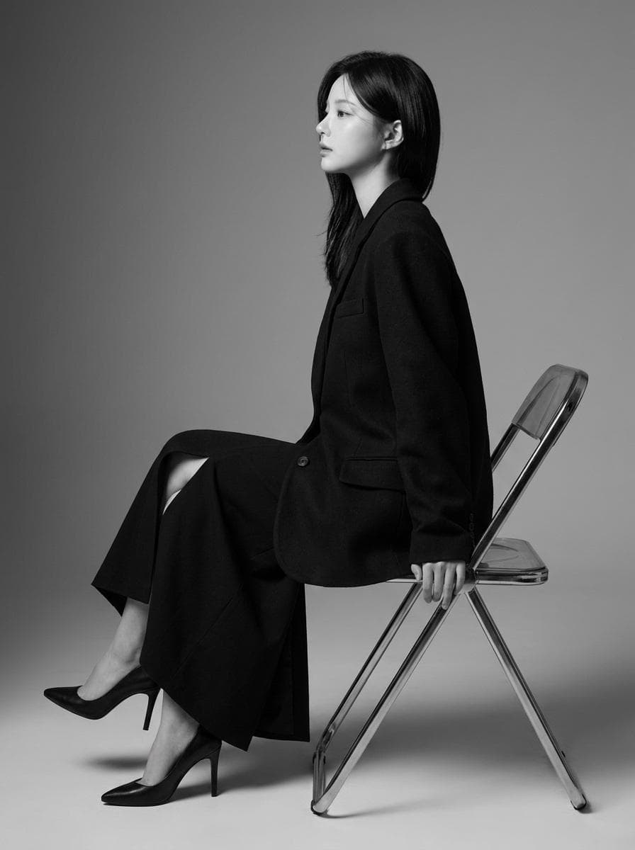 Create a highly realistic 8K professional black-and-white fashion editorial portrait of a Korean East Asian female model in her late 20s to early 30s. The model should have a serene, contemplative, and confident expression, captured in a sharp left-facing profile view while gazing off-camera. Her jaw should be relaxed, lips closed, and overall presence calm, mature, and sophisticated. The model has fair skin with neutral undertones, a smooth satin-like texture, and a natural healthy glow. Use soft studio highlights to define the bridge of the nose, cheekbones, and chin, with gentle shadows sculpting the jawline and neck. Her facial structure should feature an elegant oval face shape, refined proportions, a sharp graceful jawline, and a delicate straight nose profile. Apply a minimalist “no-makeup” makeup look emphasizing natural elegance. Use a light-to-medium coverage satin foundation, subtle upper lash-line definition, very natural mascara, softly groomed brows with a gentle arch, barely noticeable cheek contouring, and natural-toned soft matte lips. Dress the model in a high-fashion monochrome black outfit: a tailored oversized black blazer paired with a matching long flowing black skirt featuring a high thigh slit. The blazer should have structured padded shoulders, classic lapels, a deep V-neckline worn without an undershirt, single-breasted button closure, and flap pockets. The blazer fabric should appear matte and wool-blend, while the skirt should have a heavy, elegant drape. No accessories. The styling should combine masculine tailoring with feminine elegance, creating a powerful modern silhouette. Pose the model seated on a simple industrial metal folding chair, angled in a three-quarter profile toward the left. Her head remains level and fully turned into a clean left profile. Her left hand rests elegantly on the chair’s backrest while her right arm stays relaxed. Her left shoulder is slightly raised and pushed back. Her legs are crossed at the knees, creating long elegant lines, paired with sleek black high-heeled pumps. The overall body language should feel powerful, poised, statuesque, and refined. Place the subject in a minimalist professional photo studio with a seamless monochromatic gray background transitioning from medium gray to light gray. Keep the composition clean and uncluttered, allowing the strong silhouette of the model and chair to stand out. Use deep depth of…