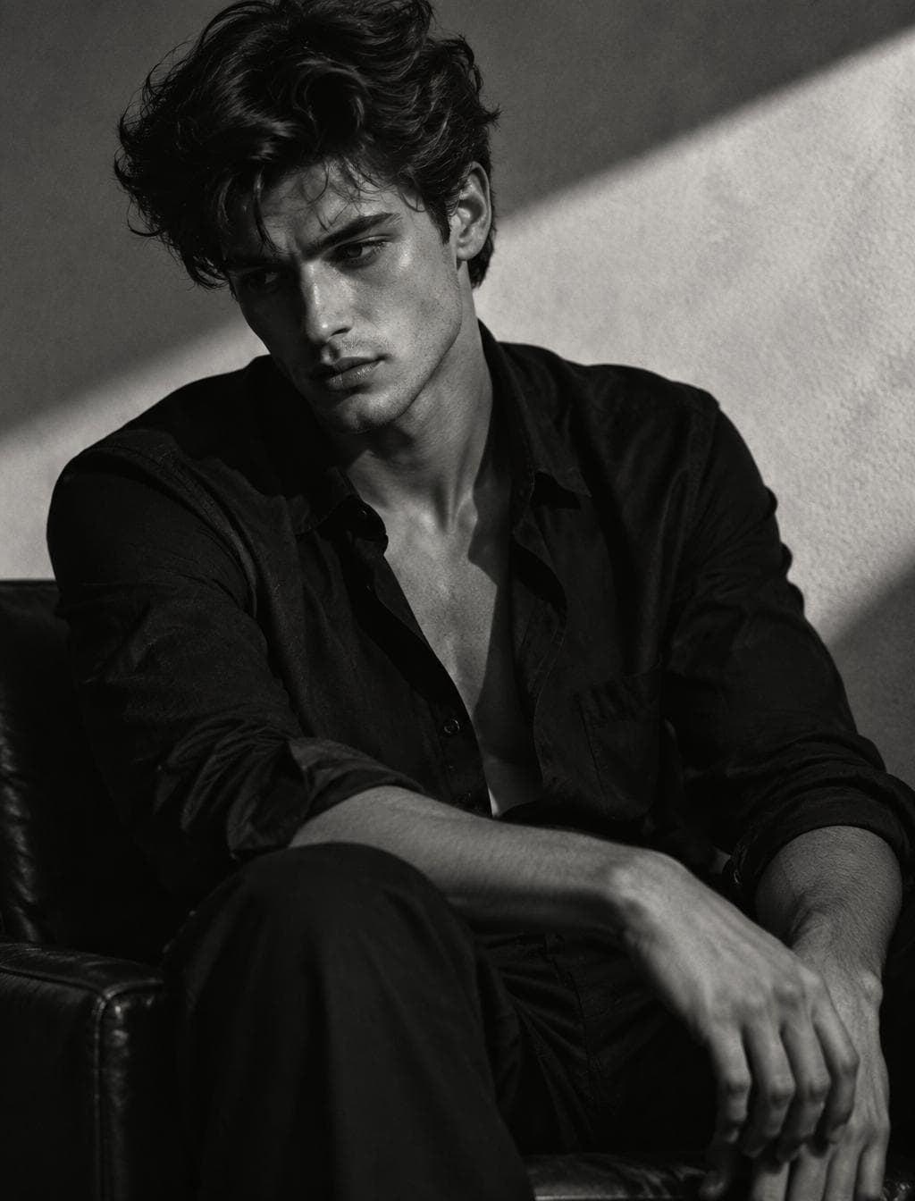 Ultra-realistic cinematic black-and-white editorial portrait, young man with thick wavy textured hair, wearing a slightly unbuttoned black dress shirt, seated in a relaxed pose, head tilted downward while looking past the camera, defined jawline, soft natural skin texture, moody expression, minimalist studio backdrop with a dramatic diagonal beam of sunlight across the wall, high-contrast Rembrandt lighting, deep noir shadows, subtle film grain, luxury fashion campaign aesthetic, photographed on a…