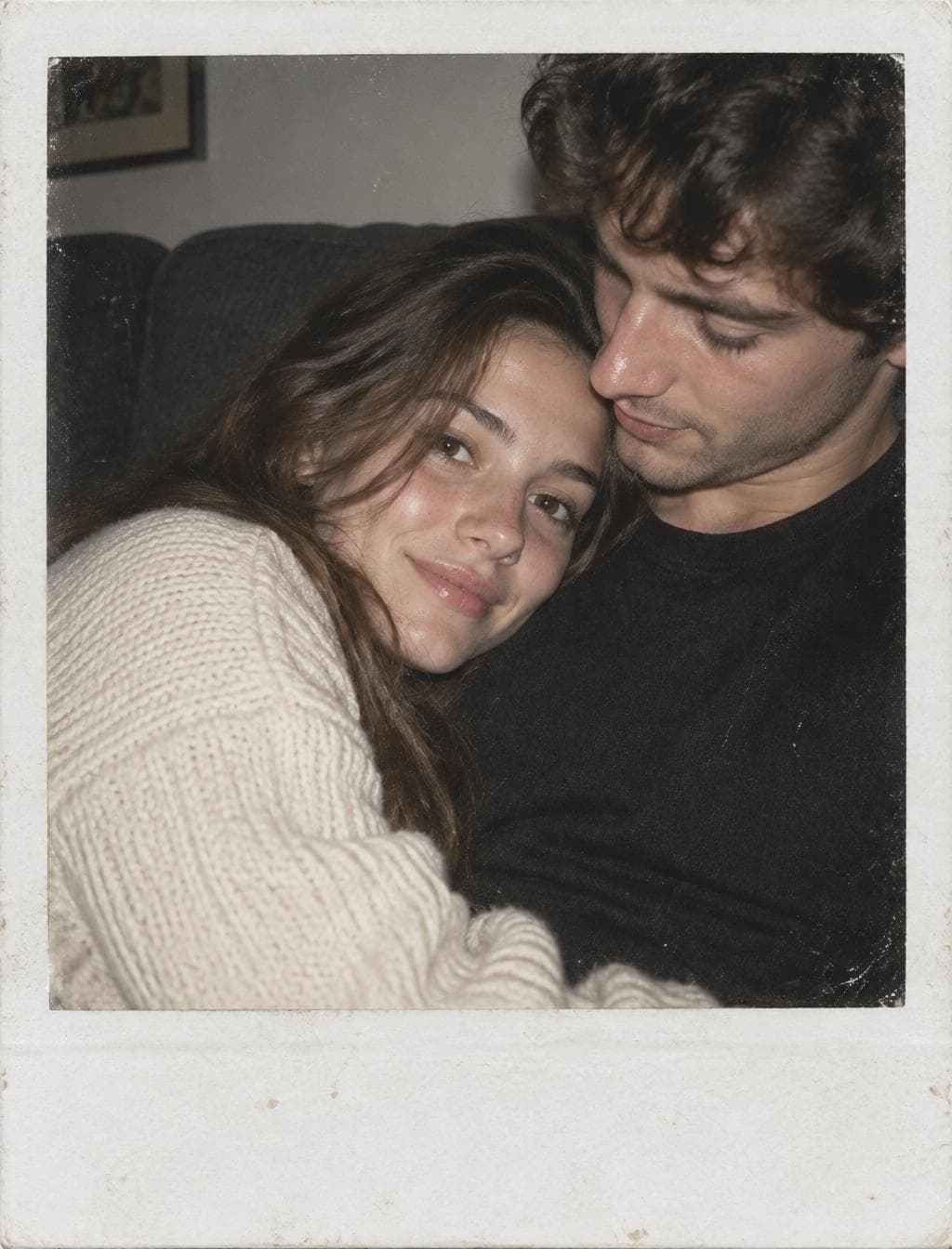 A close, intimate instant-film portrait captures a young couple reclining together, the woman leaning into the man’s chest while facing the camera with a soft, relaxed expression, and the man gazing down toward her. A direct on-camera flash creates broad frontal illumination, gentle facial shadows, glossy catchlights in their eyes, and subtle specular highlights across skin and lips. Her long, tousled brown hair and chunky cream knit sweater provide tactile softness against his matte black shirt, while a dark upholstered interior recedes behind them. The square composition is tightly cropped within a thick, slightly distressed white Polaroid border, with visible dust, scratches, muted contrast, warm color cast…