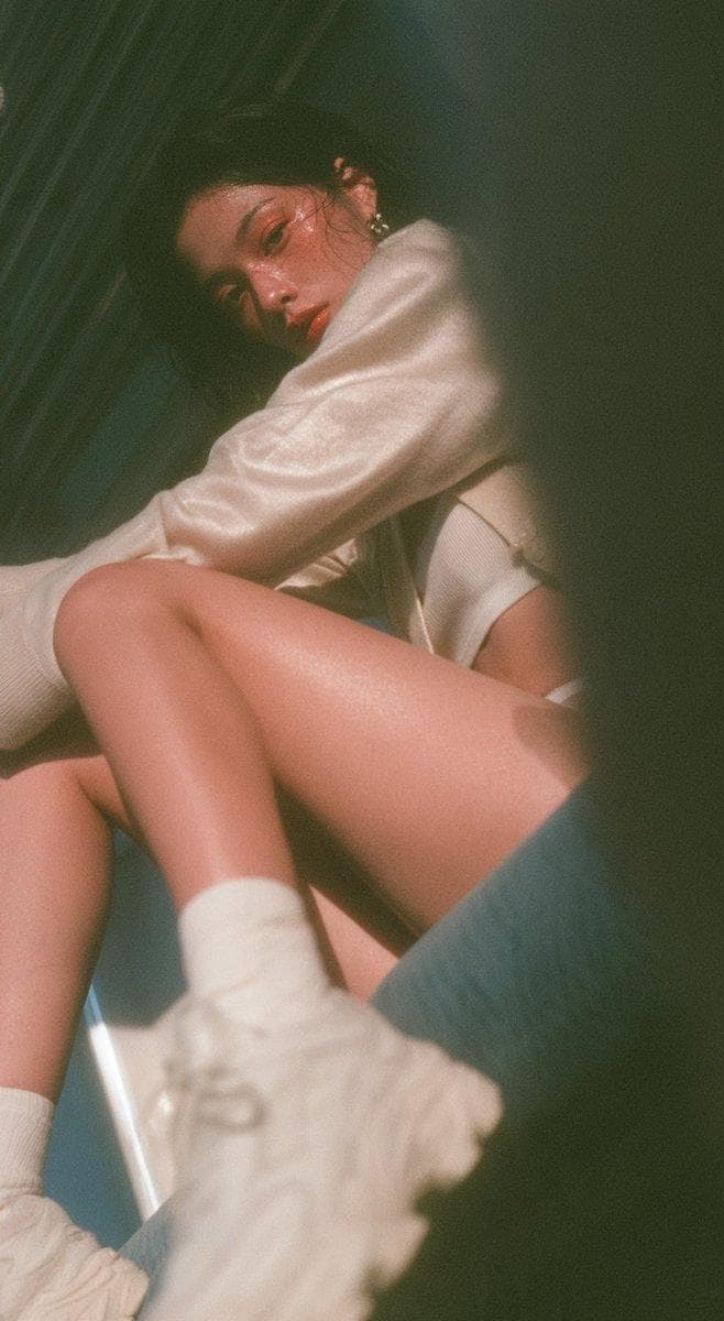 A clearly adult Taiwanese Instagram influencer, fair skin with subtle freckles, fox like refined face, softly sculpted features, pear shaped figure. Wearing a silky cashmere cardigan over a white tank top, hot pants, white sneakers. Sitting on a Tokyo subway seat in a natural, slightly dynamic pose, looking directly at camera. Foreground partially obscured for a candid snapshot feel. 9:16 vertical composition. Cool blue toned Japanese summer portrait aesthetic: low saturation, low contrast, soft haze, slight soft focus, airy atmosphere, subtle glow, gentle…