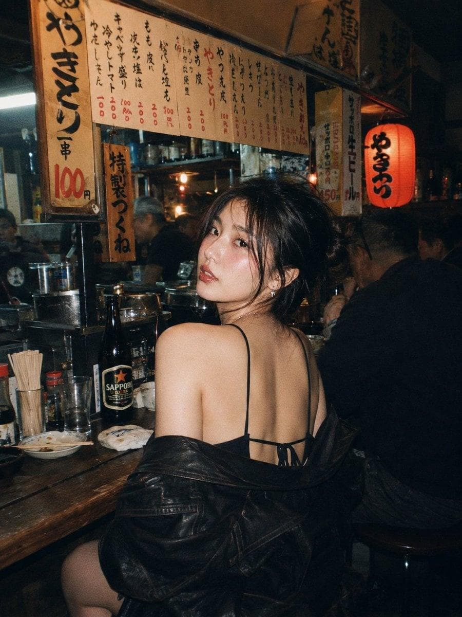 A beautiful young East Asian woman with long dark wavy hair and striking makeup, sitting at a wooden counter in a cozy, dimly lit Japanese izakaya at night. She is looking back over her shoulder directly at the viewer with a seductive gaze. She wears a white tank top under a red knitted sweater that's slipped off one shoulder, revealing her back and shoulder, paired with tight black leather shorts and sheer black stockings. The atmosphere is warm and intimate with soft lighting from a hanging lantern. Behind her are other Japanese men sitting…