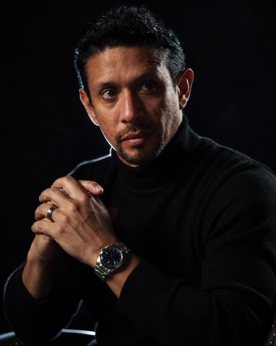 A hyper-realistic cinematic portrait of a man with short, textured hair and a well-groomed stubble beard, captured in a dark, moody 'night mode' aesthetic. The man is sitting in a contemplative pose, hands clasped together near his chest, fingers interlaced, with his gaze directed thoughtfully off-camera to the left. He is wearing a premium black high-neck turtleneck sweater and a silver-link luxury wristwatch on his left wrist, with a simple band on his right ring finger. The lighting is dramatic and controlled: a soft rim light outlines his silhouette against an almost pitch-black, shadow-drenched background, while a subtle, soft key light illuminates his face and hands. Shallow…