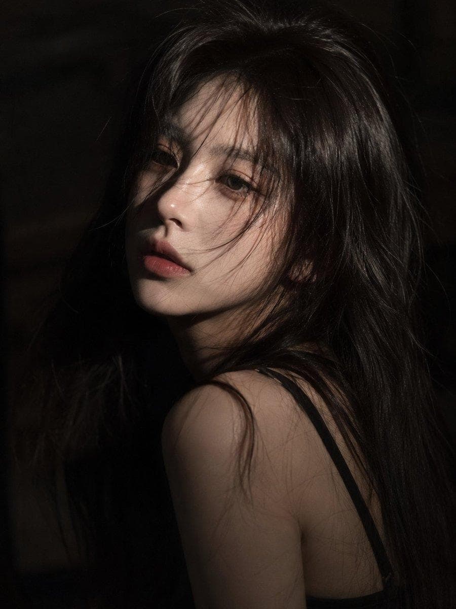 A cinematic close-up portrait of a young East Asian woman with long slightly messy black hair falling across her face, porcelain skin, soft pink lips, and deep expressive eyes. She gazes directly into the camera with a calm, melancholic expression. Dramatic low-key lighting illuminates only parts of her face, creating strong contrast against a dark shadowy background. Subtle strands of hair move gently as if touched by a breeze, adding a natural and emotional feel. Moody atmosphere…