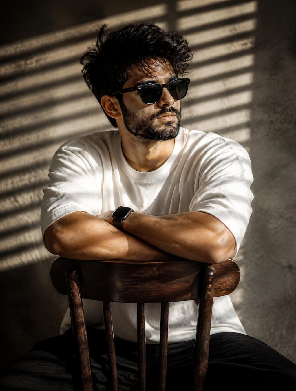 A high-resolution, cinematic portrait of a young man with messy, voluminous dark hair and a well-groomed beard. He is wearing classic black wayfarer-style sunglasses and a clean, oversized white t-shirt. The man is seated backward on a vintage dark wood chair, resting his muscular, crossed forearms comfortably along the top of the curved backrest. On his left wrist, he wears a sleek black smartwatch with a rectangular face. The scene is defined by dramatic, high-contrast lighting filtered through Venetian blinds, casting sharp, rhythmic horizontal shadow bars across his face, torso, and the textured, warm-grey plaster wall in the background. The atmosphere is moody and serene, with a shallow…