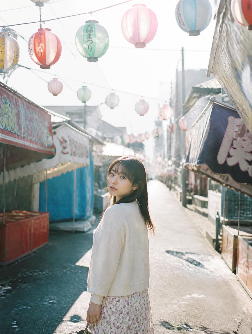 A beautiful Japanese woman walking through an empty festival street the morning after a summer matsuri, colorful lanterns still hanging overhead, abandoned food stalls, paper decorations gently moving in the breeze, wearing a simple cream cardigan over a floral dress, looking back naturally, authentic Japanese small town, Fujifilm Pro 400H, Nikon FM2, soft overexposed morning sunlight, pastel color palette…
