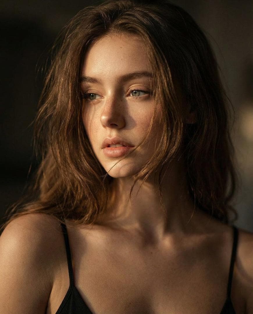 Ultra-realistic Cinematic Close-up Portrait, Replace Subject With My Reference Image. A Young Woman Shown From Shoulders Up, Head Slightly Turned, Eyes Looking Off-frame With A Soft. Thoughtful Expression. Natural Facial Proportions, Visible Skin Texture, Subtle Freckles And Fine Details Preserved. Loose, Slightly Messy Hair Strands Falling Across The Face, Creating A Natural, Intimate Mood, Wearing A Thin-strap Black Top, Minimal Styling, No Heavy Accessories. Lighting Is Dramatic And Cinematic: Warm Directional Light Hitting The Face From One Side, Deep Shadows On The Opposite Side, Creating Strong Contrast And Emotional Depth. Background Is Dark And Unobtrusive, Softly Blurred, Keeping Full Focus On The Face. Shot With A…