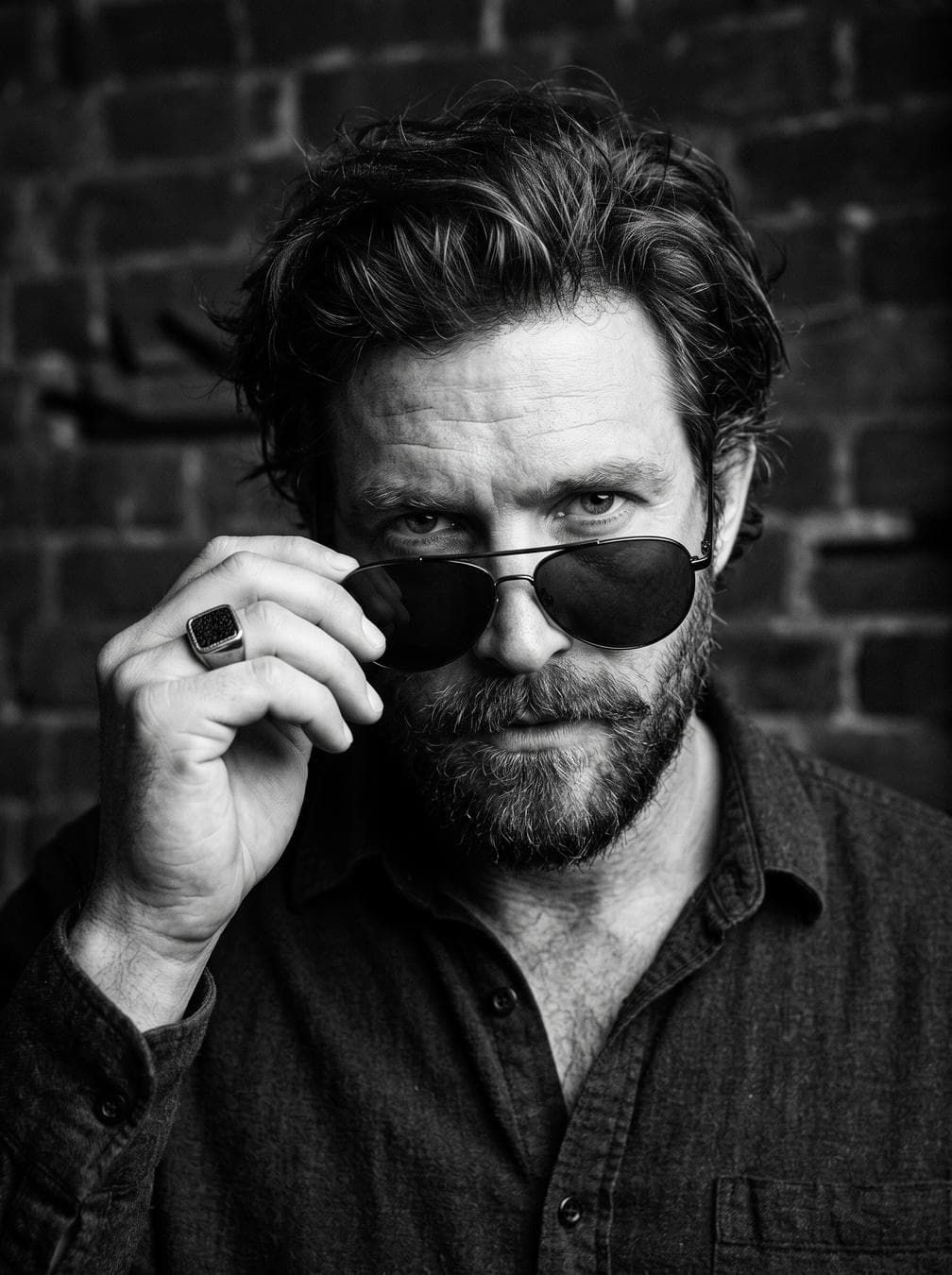 A close-up, high-contrast black and white portrait of a man looking directly into the camera with an intense expression. He has thick, voluminous messy dark hair, a neatly trimmed beard, and a mustache. He is wearing dark aviator-style sunglasses with a double bridge bar, which he lowers slightly down his nose with his right hand, peering directly over the top rims. On the ring finger of his right hand, he wears a prominent square signet ring with a dark center inlay. He is dressed in a dark button-up shirt, left open at the collar to reveal a hint of his chest. The…