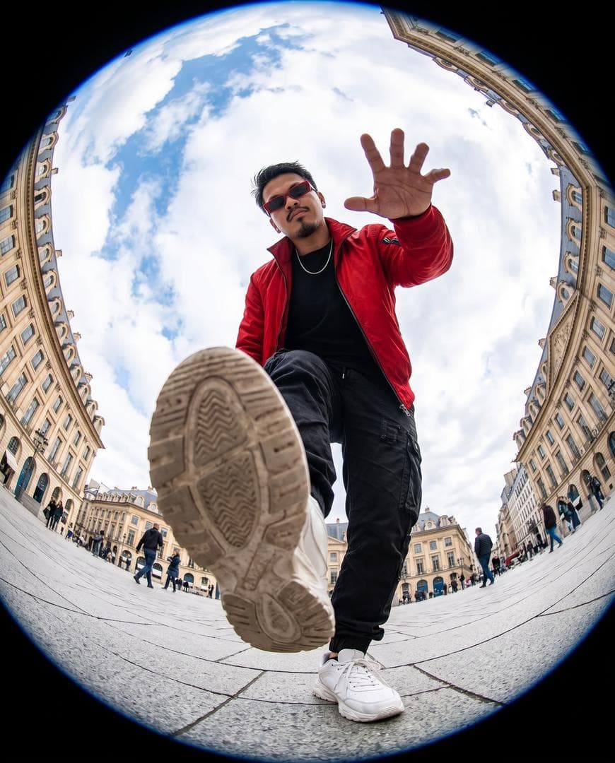 Ultra-realistic action photography, extreme fisheye lens, 8mm, worm’s-eye view from ground level, dynamic urban plaza scene. A stylish young person appears to be stepping over the camera, one sneaker sole dominating the foreground, hand reaching toward the lens.. Massive circular “tiny planet” perspective with historic European architecture curving around the edges of the frame, bright blue sky filled with fluffy white clouds at the center. Cinematic composition, dramatic distortion, ultra-wide angle, vibrant…