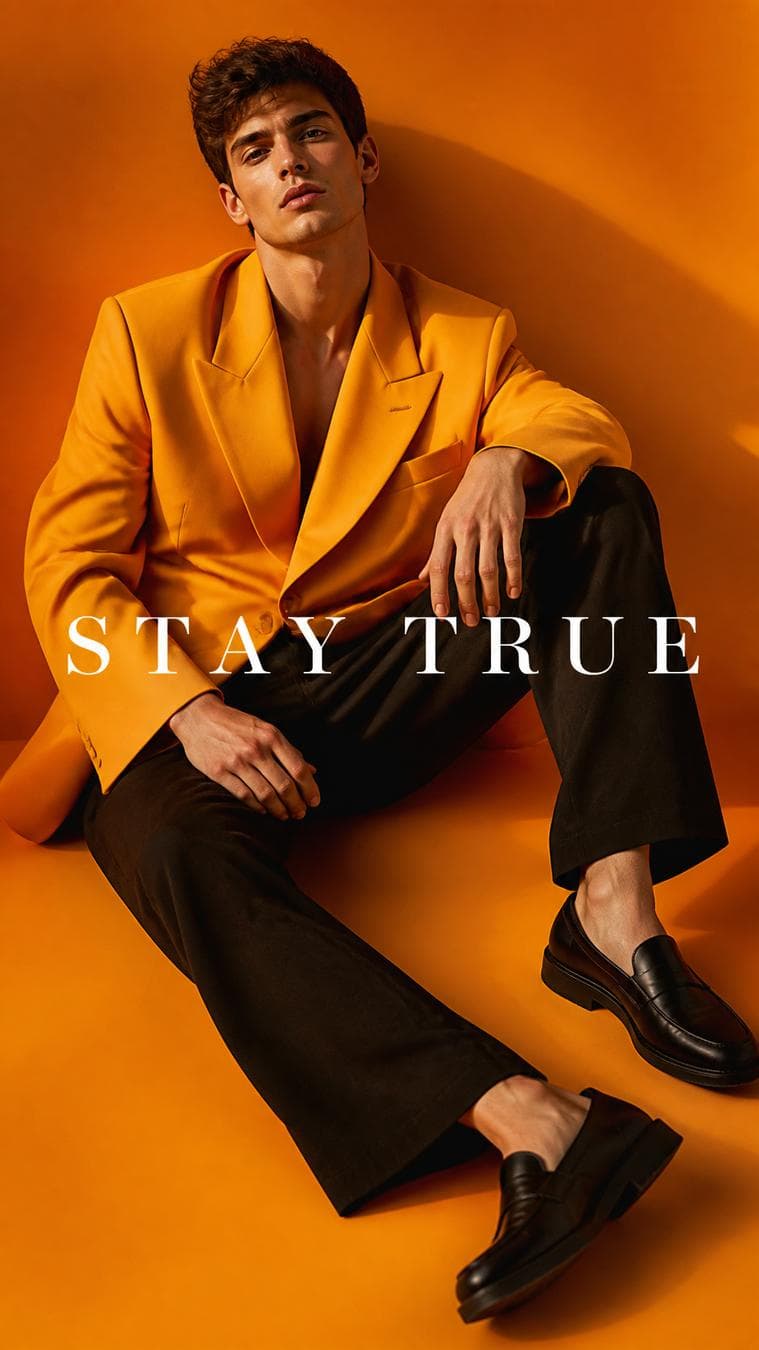 Luxury fashion campaign. Main Subject: Male model wearing a bright saffron oversized blazer. Pose: Sitting casually on the floor with one knee raised. Camera: Slight overhead perspective. Background…