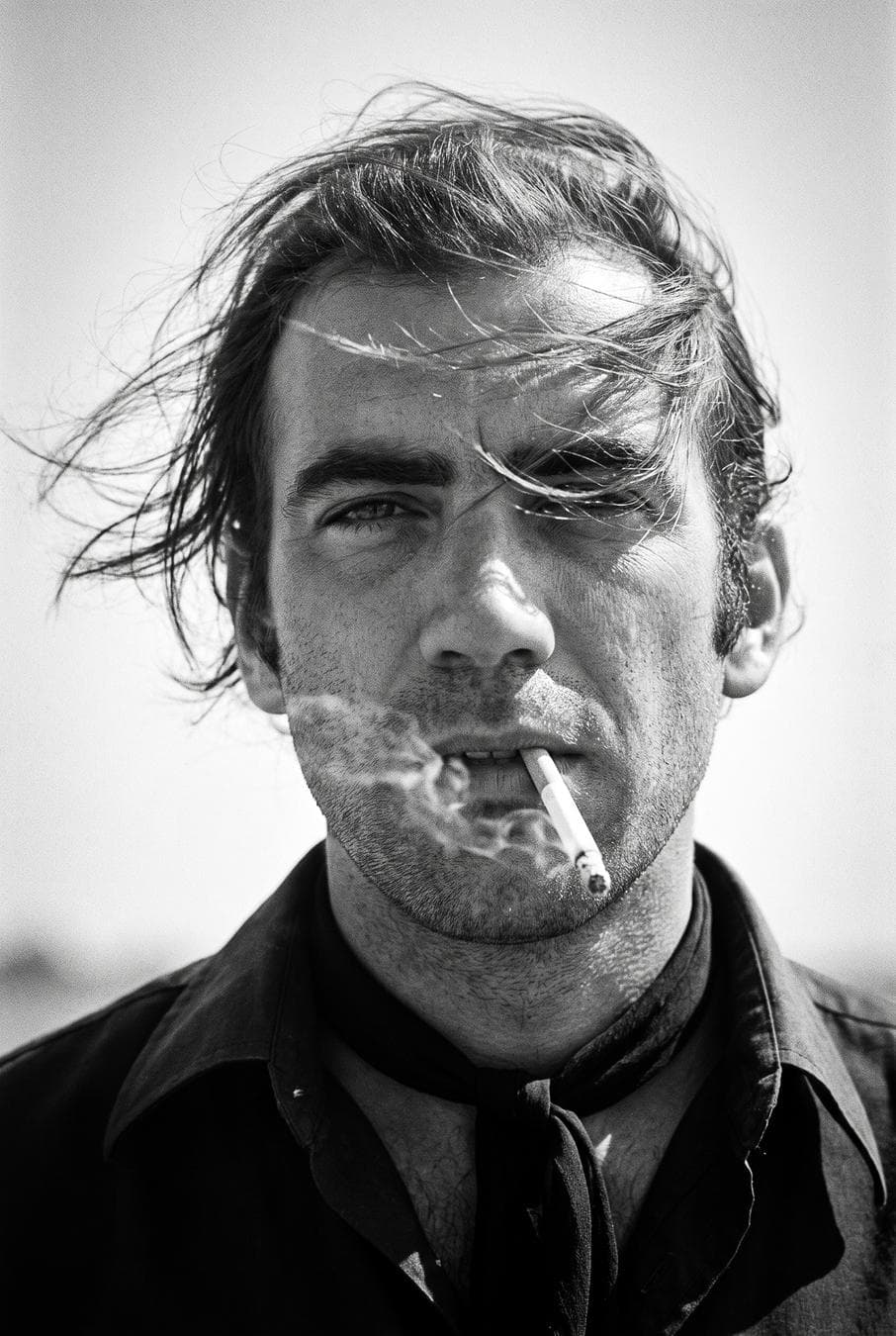 Create a 1970s black-and-white close-up portrait in a windswept outdoor setting, black and white noise.Camera/look: eye-level, front ¾ view (~30° off-axis); 85–105 mm portrait perspective; vertical 2:3, tight head-and-shoulders crop; shallow DoF f/2.8–f/4; subject centered.Lighting: hard natural sunlight from upper camera-left with minimal fill; crisp shadows; subtle rim on hair; sky as bright mid-tone background; no blown highlights.Pose & expression: subject faces camera, chin slightly down, lips parted with a cigarette held between them; gaze relaxed/half-lidded; hair blown across the face by wind, partially covering one eye.Wardrobe & styling: dark collared shirt with open neck; thin black neck scarf tied loosely; no logos/jewelry.Grade: high-contrast monochrome with rich…