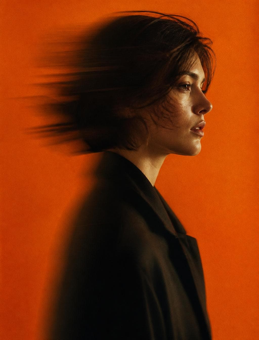 { "image_generation": { "subject": { "description": "mysterious young woman portrait", "pose": "side profile portrait with head slightly turned forward", "expression": "calm, serious, mysterious mood", "gaze": "looking forward into the distance" }, "appearance": { "hair": { "color": "dark brown", "style": "short, wind-blown hair with motion blur" }, "skin": { "tone": "soft natural tone", "texture": "smooth cinematic skin texture" } }, "outfit": { "top": "dark minimal coat or jacket with high collar" }, "environment": { "background": "solid orange studio background", "setting": "minimal studio environment with clean backdrop" }, "lighting": { "type": "studio lighting", "style": "soft diffused light with cinematic contrast", "effect": "subtle glow and smooth shadow gradients" }, "camera": { "angle": "side profile portrait shot", "framing": "medium portrait", "effect": "panning motion blur creating dynamic movement" }, "aesthetic": {…