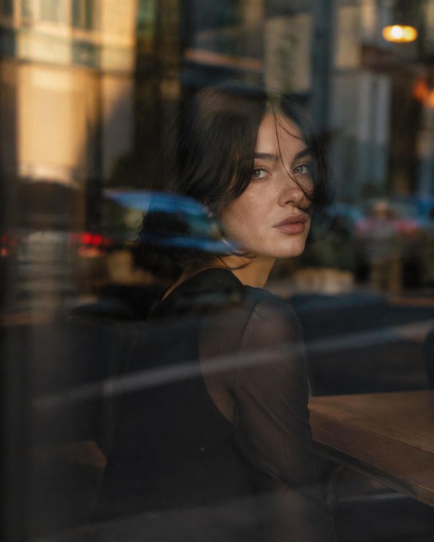 Ultra-cinematic close-up portrait of a young woman sitting inside a dark urban café, photographed from outside through a large reflective glass window. She has a sleek chin-length black bob with a center part, soft loose strands falling naturally across her face, pale realistic skin with subtle freckles, light green-gray eyes, soft natural lips, minimal makeup, and a quiet, distant, melancholic expression. She wears an elegant all-black outfit with sheer black sleeves. Her face is visible beneath multiple layers of glass reflections: blue streaks from passing cars, blurred city buildings, warm orange lights, street silhouettes and abstract urban shapes flowing across the frame. The reflections partially obscure her face, creating a dreamy double-exposure effect. Warm golden-hour sunlight gently cuts across sections of her face and glossy hair while the café interior remains dark and atmospheric. Intimate candid composition, subject looking sideways or subtly toward the camera, natural unposed body language, shallow…