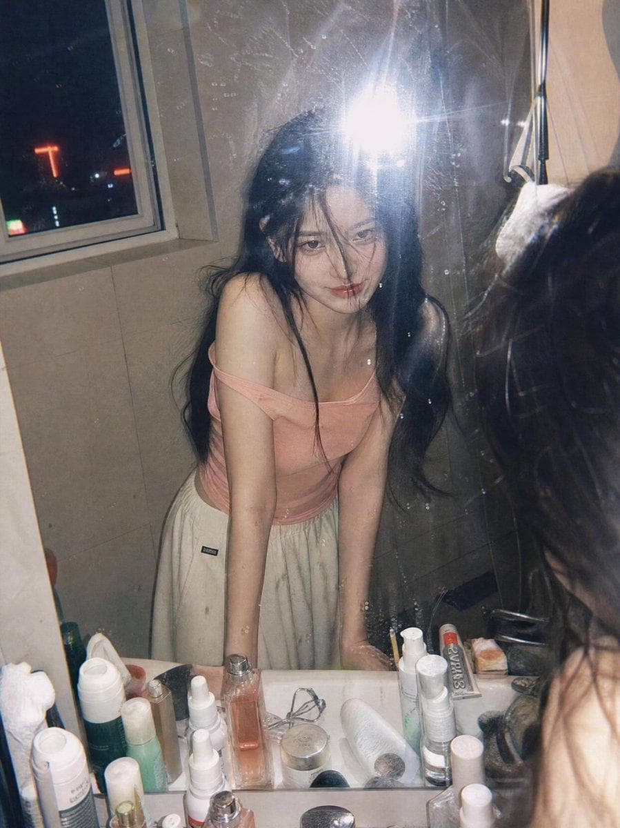 Ultra-realistic candid film photography portrait of a young woman leaning over a bathroom sink and looking into a mirror at night, captured through a slightly dirty mirror with natural reflections and subtle imperfections. She has long black hair partially covering her face, soft glowing skin, and a gentle mysterious smile. Wearing a light pink off-shoulder fitted top and loose cream-colored lounge pants, she poses casually with both hands resting on the sink. The scene is illuminated by direct on-camera flash, creating authentic Y2K disposable camera aesthetics, soft highlights, realistic skin texture, and natural shadows. A nighttime city skyline with colorful neon lights and high-rise buildings is visible through a nearby window, adding an urban atmosphere. The bathroom countertop is filled with skincare and cosmetic products, enhancing the lived-in, personal feel. Shot from a close-up mirror…