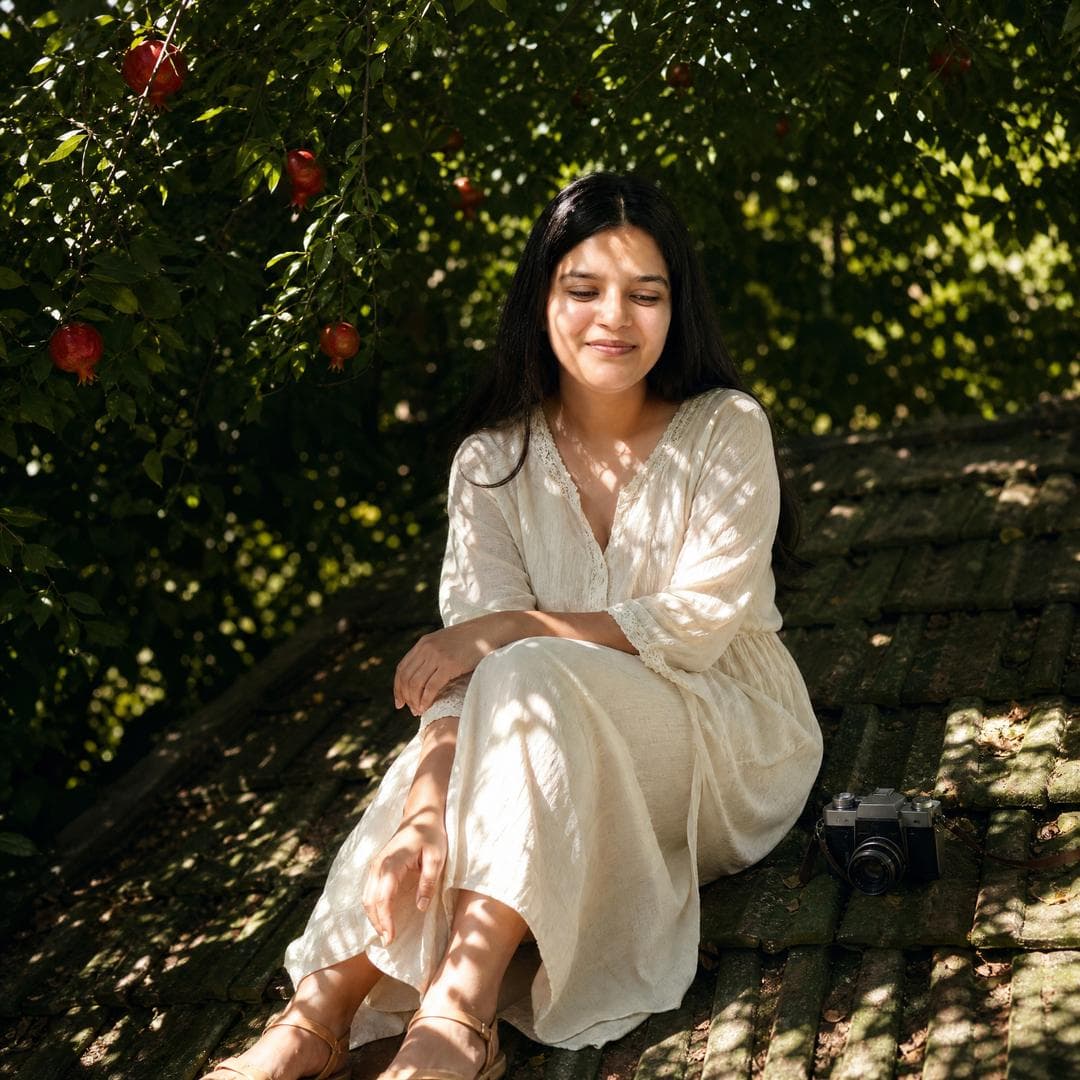 Use the uploaded portrait only as the identity reference. Preserve the person’s real facial features, face shape, skin tone, and recognizable hairstyle. Do not copy the original outfit, background, pose, lighting, or image quality from the uploaded portrait. Create a realistic cinematic portrait of the same person sitting quietly on an old green-tiled rooftop under dense tree shadows. The person is relaxed, looking slightly downward with a soft calm expression. Surround the scene with dark green foliage, hanging red pomegranate fruits, scattered sunlight, and natural leaf-shaped shadows across the face, body, and roof tiles. Add a vintage handheld camera placed casually nearby as a storytelling detail. Choose an outfit that best matches the person’s appearance and personality, different from the scene inspiration: natural, clean, elegant, lightweight, and suitable for a dreamy outdoor summer portrait. Keep the styling tasteful and realistic. Low-angle composition, full-body or three-quarter body framing, cinematic natural light, strong contrast between deep shadow and bright sun patches, soft film grain, subtle lens flare, dreamy atmosphere, realistic skin texture, natural body proportions, authentic photography, shallow depth of field, poetic summer mood…