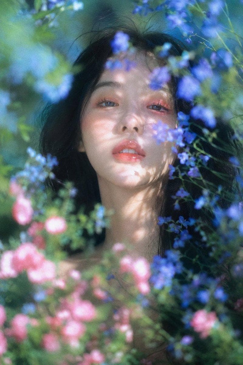 梦幻自然光花园人像,柔焦胶片质感,竖幅近距离肖像构图;年轻女性,muse般的面庞,黑色长发蓬松微乱,面部居中偏上作为主视觉,白皙偏暖象牙肤调,橘粉眼妆与水润珊瑚唇,正面安静凝视镜头,头部略抬,嘴唇轻启,眼神湿润疏离、无笑意;身体只露出少量肩颈,被花枝遮挡。前景粉色、蓝紫色小花和绿色枝叶中等密度环绕面部并局部遮挡,背景蓝绿色自然虚化…