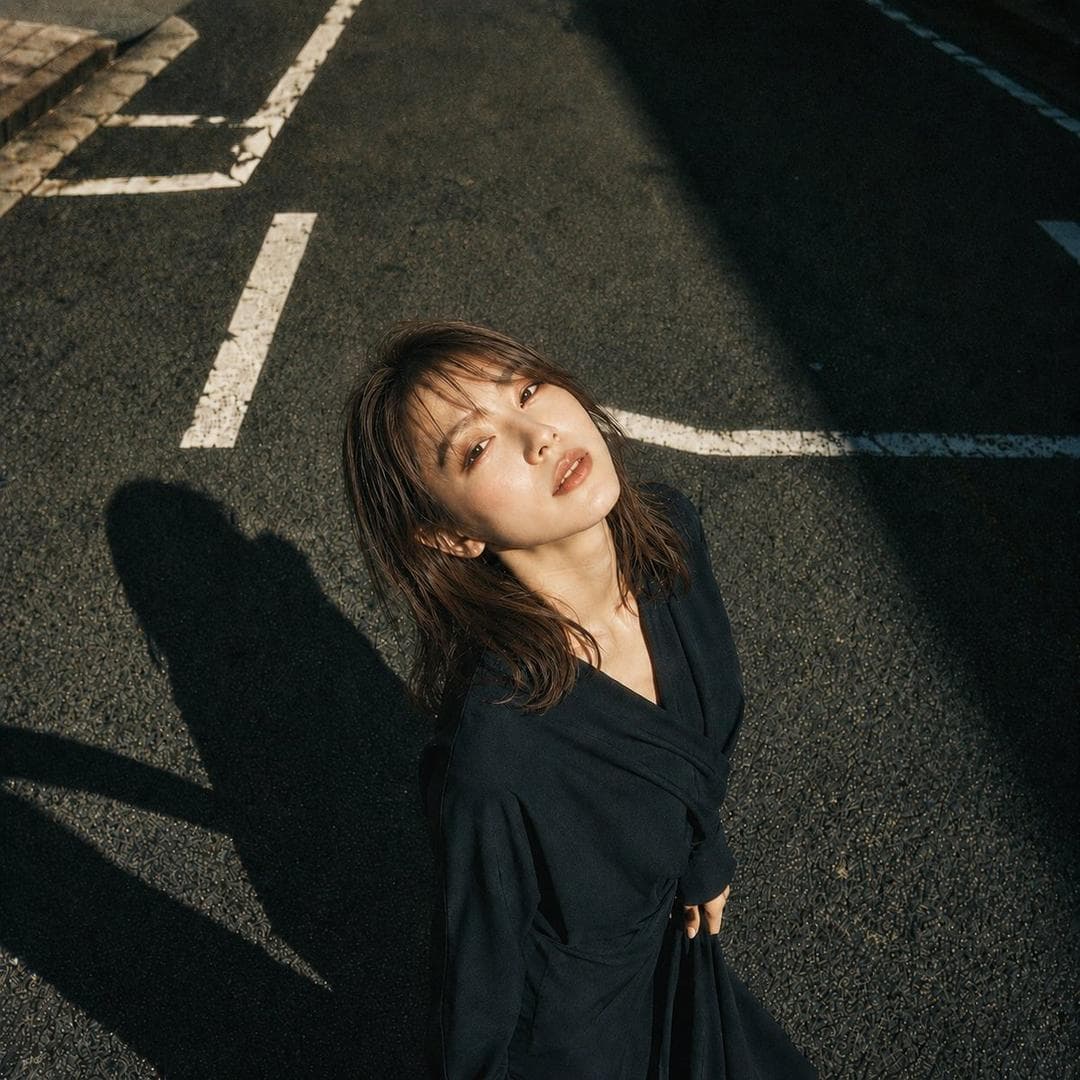 主題:白線と影 人物・表情: 20代女性。明るいアイボリーベージュ寄りの白肌に、ほんのりピンクの血色を含んだ自然な肌質。頬、鼻先、鎖骨に柔らかなハイライト。精密で上品なドールライク日系メイク、淡いローズピンクのチーク、シャンパンベージュとピンクブラウンの目元、細く長いまつ毛、自然な涙袋、明るめブラウンの柔らかい眉、透け感のあるピンクベージュのグロスリップ。瞳に小さなキャッチライト。静かに見上げる、少し余韻のある表情。 服装・ポーズ: 深い黒に近いネイビーの和モダンなラップワンピース。長袖、重なりのあるVライン、腰で布を結んだようなドレープ、落ち感のある柔らかな生地。身体は画面下側に立ち、上半身を少し後ろへ預ける。顔はカメラへ向けて上げ、右手はスカートの布を軽く持ち、左腕は自然に体側へ落とす。姿勢は大人っぽく、静かな緊張感を残す。 背景・光: 日本の都市のアスファルト道路。白い道路標示が斜めに入り、画面左上に歩道の縁石が少し見える。強い午後の太陽が画面左上から差し、顔と髪、胸元を明るく照らす。人物の影は画面左下へ長く伸び、画面右上には建物の濃い影が大きく落ちる。白線、影、アスファルトの粒感をはっきり描写する。 構図・カメラ:…