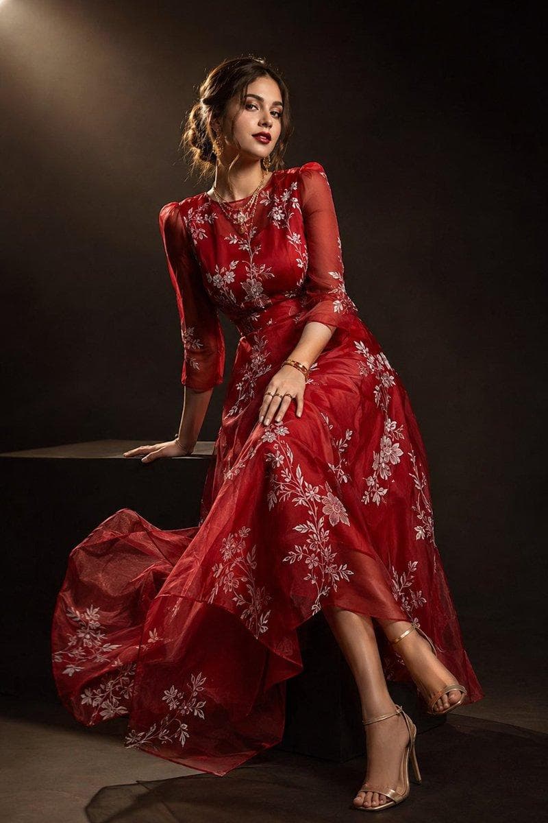 Ultra-realistic high-fashion Vogue magazine editorial portrait of a beautiful young woman wearing an elegant deep ruby-red organza gown crafted from luxurious sheer layered fabric. The gown features a modest sophisticated neckline, graceful 3/4-length sheer sleeves, a fitted waist, and a flowing A-line skirt extending below the knees. The entire gown is adorned with delicate artistic white floral and botanical hand-painted patterns, creating a dreamy couture aesthetic. The lightweight organza fabric catches the light beautifully with soft translucent layers and elegant movement. She accessorizes with subtle yet luxurious gold jewelry: stylish modern gold drop earrings, layered delicate gold neck chains, elegant stacked bracelets, and refined gold finger rings. Footwear consists of sophisticated nude-gold strappy heels with a polished designer look. Hair styled in a soft romantic low bun with loose face-framing strands, smooth texture, natural volume, and effortless elegance. Makeup features flawless radiant skin, peach-glow complexion, soft contouring, subtle champagne highlighter, ultra-defined arched eyebrows, long thick curled eyelashes, soft winged eyeliner, warm eyeshadow blend, and deep berry glossy lips. Pose: elegant Vogue-inspired seated fashion pose on a black studio pedestal, one leg slightly extended, shoulders angled gracefully, chin slightly lifted, confident yet feminine expression, hands delicately positioned to…