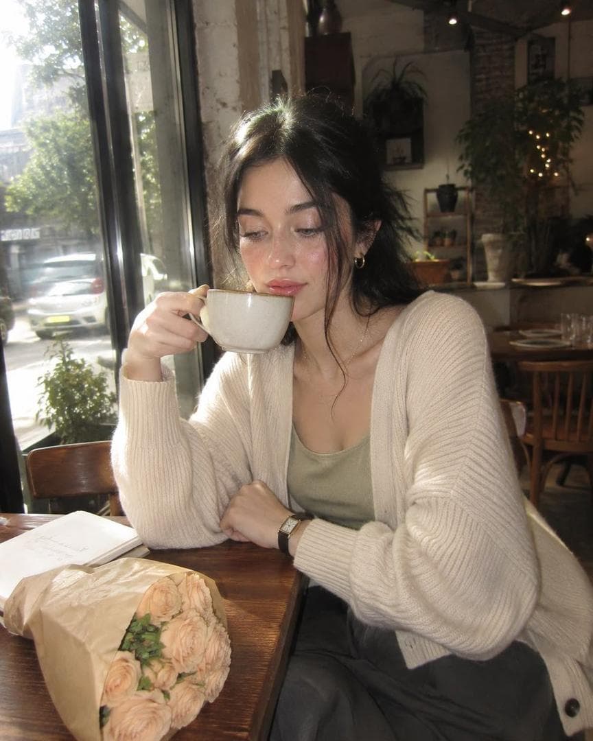 Create an ultra-realistic nostalgic café portrait captured on a premium early-2000s compact digital camera, blending direct flash with soft afternoon daylight. The image has a dreamy, imperfect snapshot quality with gentle motion softness caused by natural handheld camera movement, subtle subject motion, and shifting strands of hair. The atmosphere feels candid, intimate, and effortlessly authentic. A young woman sits alone at a cozy café table beside a large sunlit window. Instead of facing outward, she is turned slightly toward the camera while lowering her gaze with a quiet, thoughtful expression. One hand loosely holds a ceramic coffee cup near her lips while the other rests against the edge of the table. Her posture is relaxed and natural, conveying a calm afternoon moment rather than a posed portrait. She wears an oversized cream-colored knit cardigan layered over a soft sage-green top, loose charcoal trousers, a slim vintage wristwatch, and understated jewelry. On the table are a cappuccino with delicate foam art, a small notebook, and a bundle of pale peach roses wrapped in kraft paper. Warm wooden furniture, softly blurred indoor plants, scattered reflections, and muted café décor create a peaceful everyday setting. Shot through thick vintage optics with pronounced spherical aberration, reduced edge sharpness, soft focus drift, blooming highlights, subtle lens haze, gentle veiling flare, lifted blacks, low micro-contrast, and characteristic CCD digital-camera color rendering. Fine JPEG softness and natural optical imperfections contribute to the nostalgic aesthetic. Small naturally clipped highlights appear on hair strands, glossy lips, watch crystal, coffee cup edges, flower petals, table surfaces, window reflections, and bright outdoor areas beyond the glass. Light softly blooms into surrounding tones without artificial sparkle effects or exaggerated glow. Skin appears naturally healthy and luminous with realistic texture and faintly visible pores beneath the dreamy softness. Gentle warmth is concentrated in the cheeks and lips, while the complexion retains realistic tonal variation and depth. Majeup features refined glass-skin beauty styling: softly lifted brows, subtle grey-blue eyes, delicately separated lashes, muted taupe eyeshadow, and luminous reflective highlighter on the cheekbones, nose bridge, inner eye corners, cupid’s bow, and chin. The highlights remain bright and elegant while preserving believable skin texture. Colors remain rich and distinct despite the soft contrast: cream…