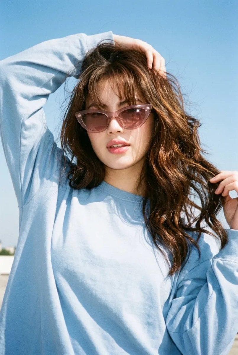 Ultra-realistic vintage Y2K summer fashion portrait of an adult woman, vertical 2:3, captured with a nostalgic 2000s point-and-shoot film camera aesthetic. She has long, thick, naturally voluminous dark chocolate-brown hair with subtle espresso and warm chestnut highlights, soft airy bangs, slightly messy face-framing strands and natural movement from the breeze. Her hair looks glossy, dimensional and effortlessly cool rather than overly styled. She wears oversized translucent dusty-pink cat-eye sunglasses with softly reflective lenses and a relaxed powder-blue oversized long-sleeve top, slightly loose and casually draped for an effortless feminine street-style look. Soft glam makeup with luminous natural skin, peachy-pink blush, softly sculpted cheeks, defined warm-brown eyes, wispy lashes, subtle brown liner and glossy rosy-pink lips. Fresh, youthful, polished but still natural. She stands outdoors beneath a clear blue summer sky. Her body is angled slightly sideways instead of facing the camera directly. One arm is lifted above her head, fingers casually touching the top of her hair, while her other hand lightly gathers a few strands of hair near her shoulder. Her head tilts naturally toward the raised arm, chin slightly lifted, lips softly parted, giving a confident, playful, effortlessly cool expression. Dynamic…